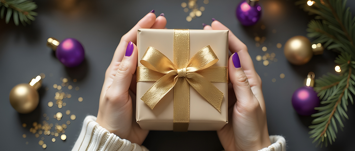 Hands with purple manicure holding a gold-ribboned gift box among holiday ornaments, for salon holiday retail tips