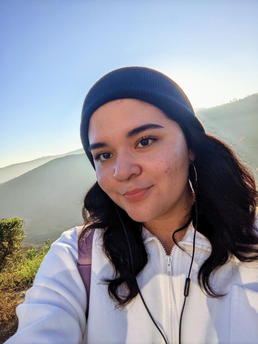 A light-skinned person with long dark brown wavy hair, wearing a beanie. They are standing in front of a bright sky.