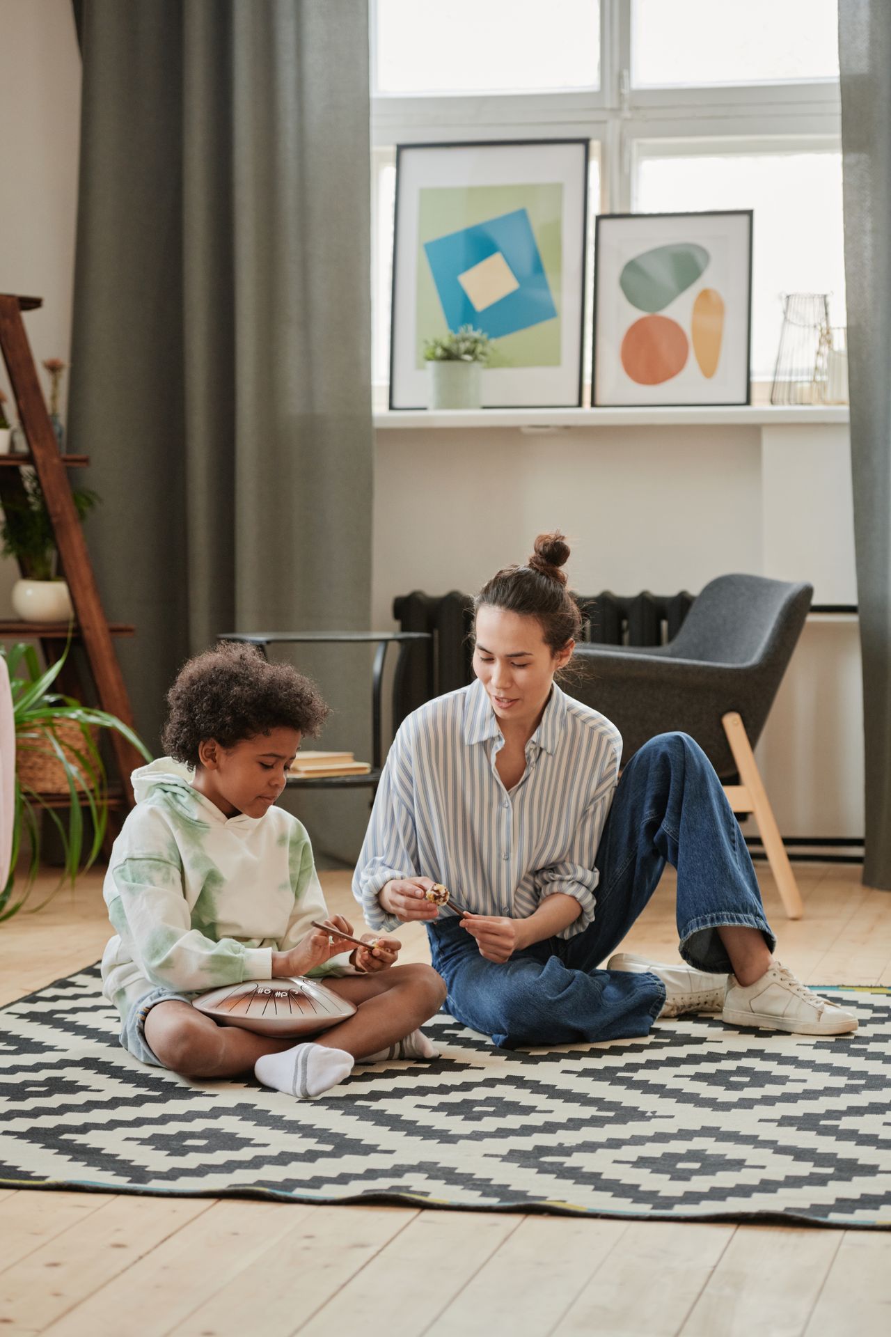 A child and an adult sitting on a patterned rug in a cozy room; the child plays a steel tongue drum while the adult watches and holds a mallet, with modern art prints and a chair in the background.