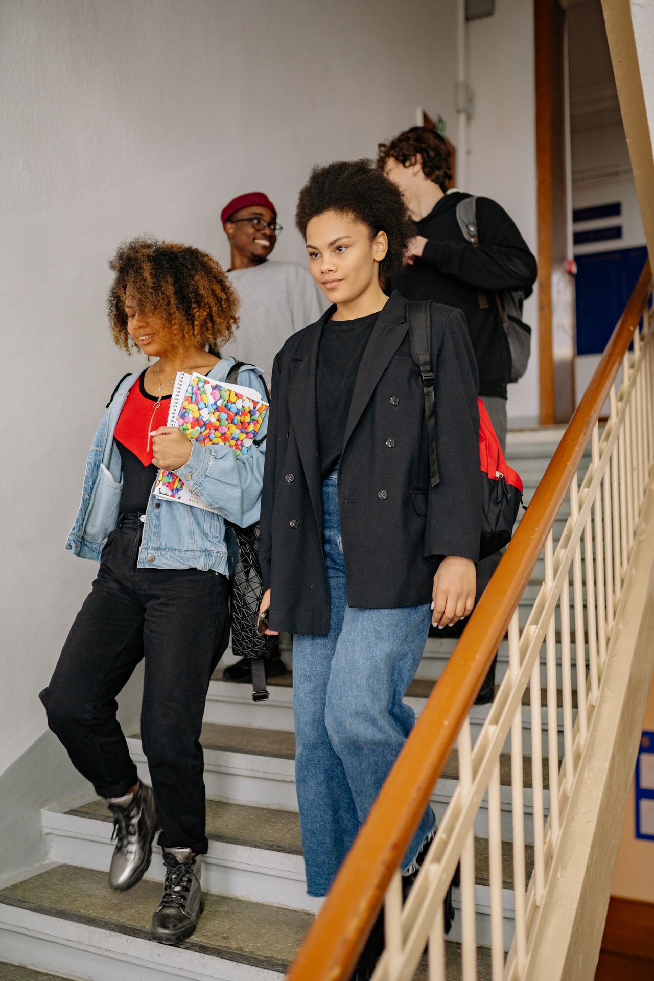 Four diverse teenagers walking down indoor stairs, two girls in front holding notebooks and backpacks, two boys behind them chatting and smiling.