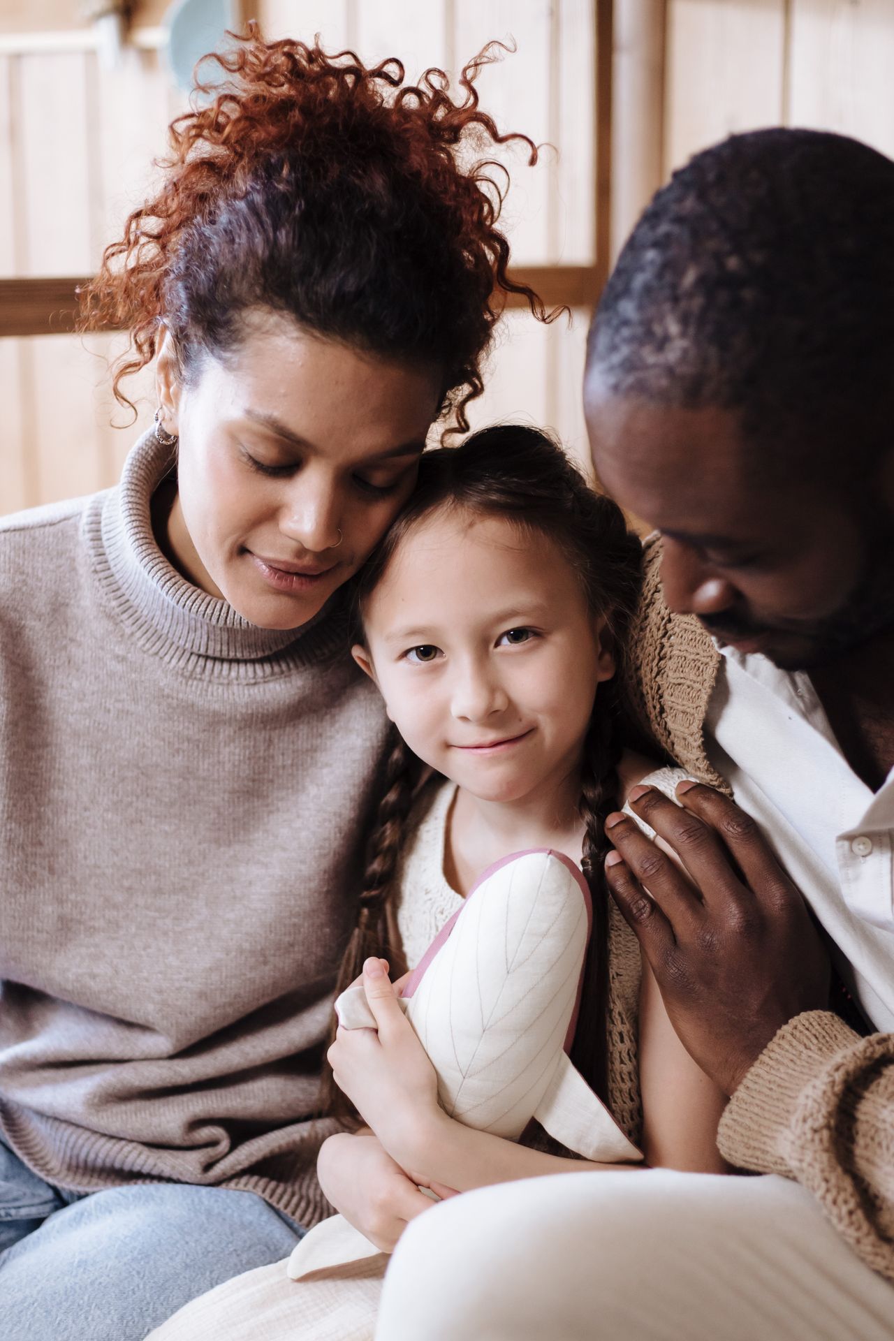 A young girl with braided hair holding a white plush toy, sitting between two adults who are leaning close in a warm, tender family moment indoors.