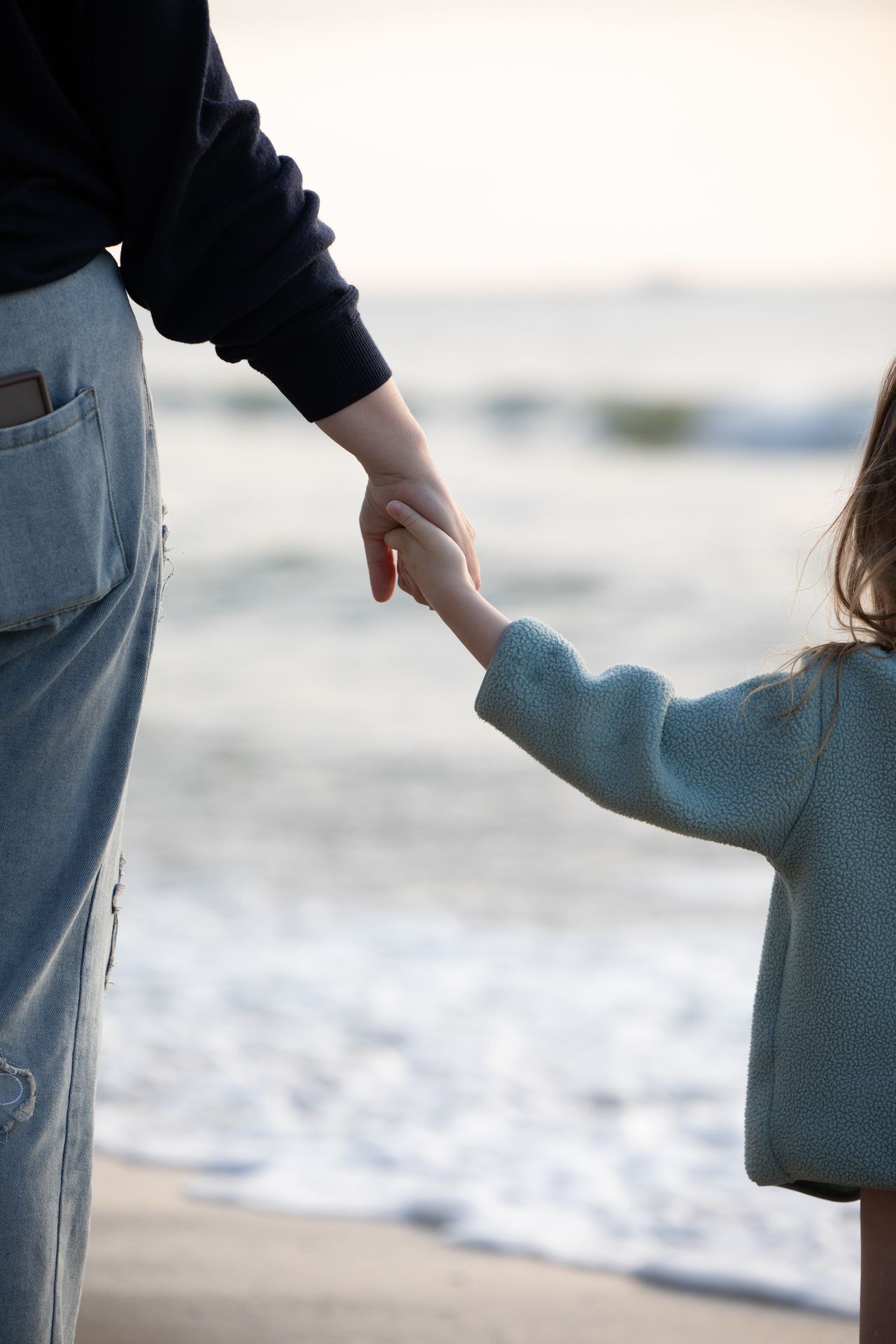 Adult and child holding hands on a beach near the water's edge, with the child wearing a light blue jacket and the adult in a dark sweater and jeans.