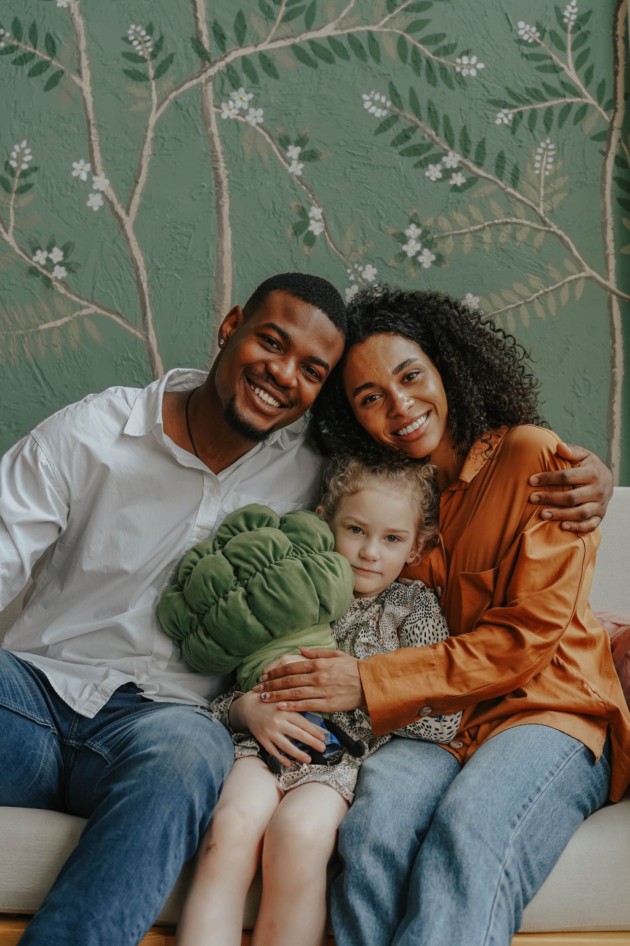 Smiling couple hugging a young child sitting between them on a sofa, with a green wall featuring a floral branch pattern in the background.