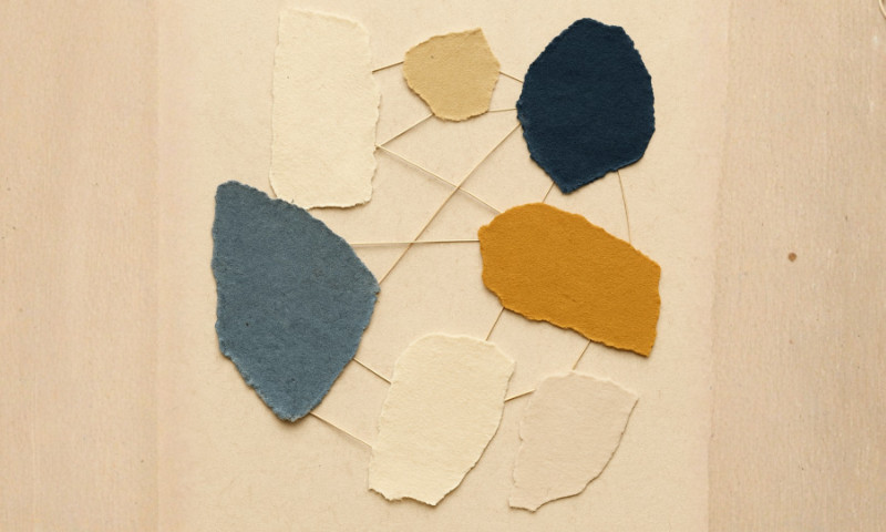 A minimalist torn-paper collage showing irregular paper fragments in muted blue, ochre, and cream tones, connected by thin thread lines in a loose circular pattern, symbolising collaboration and team connection.