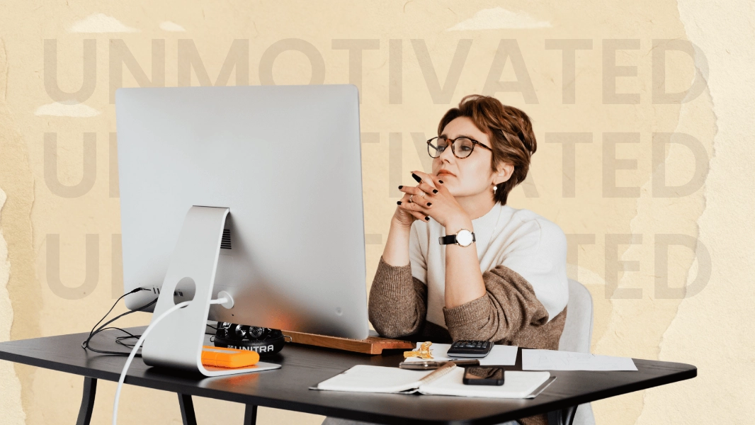 A woman sits at a modern desk, leaning forward with her hands clasped under her chin, staring thoughtfully at a large desktop monitor. She wears glasses, a watch, and a neutral-toned sweater layered over a white top. Papers, a phone, pens, and a small snack sit scattered around her workspace, suggesting a moment of fatigue or low motivation. Behind her, the textured beige background includes faint, oversized, semi-transparent lettering that repeats the word “UNMOTIVATED,” reinforcing the mood. The overall scene conveys someone feeling stuck, disengaged, or unsure of what to do next.