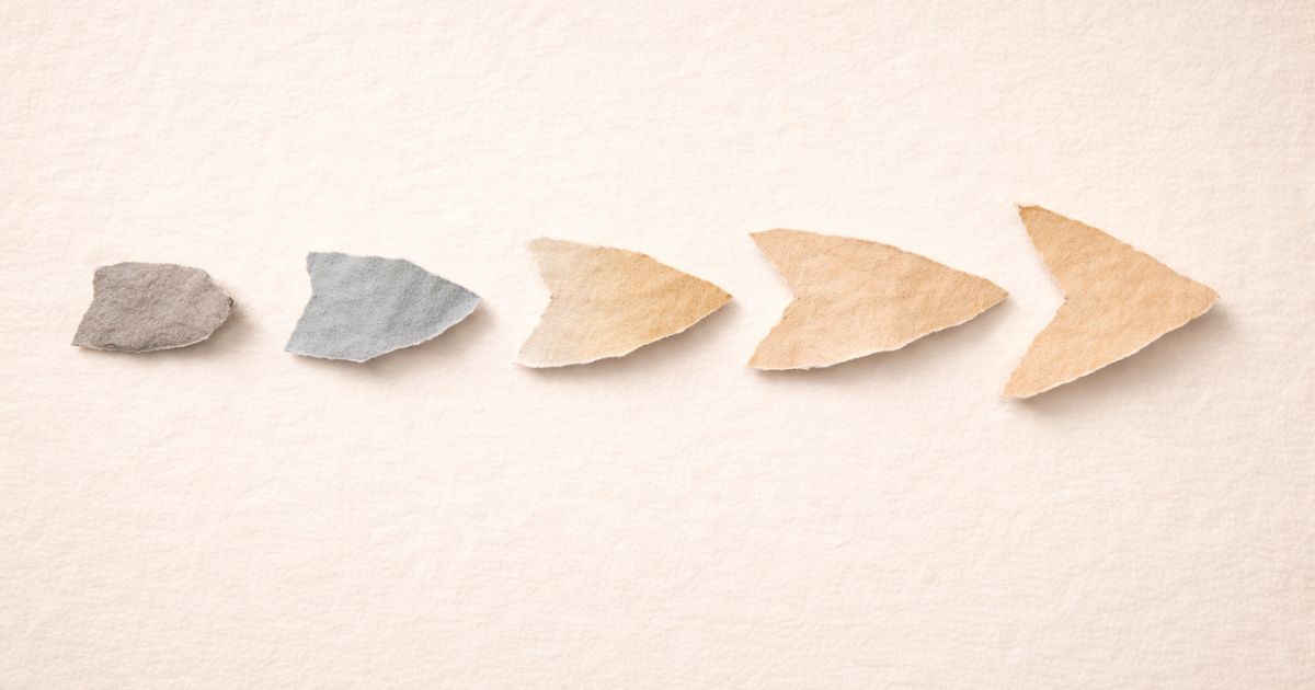 Minimalist torn-paper collage on a warm cream textured background showing a horizontal progression of five hand-torn paper shapes moving from left to right. The first shape on the far left is small, compact, and irregular in a muted stone grey tone, with rough fibrous edges and a soft shadow giving it slight lift. As the sequence continues, each shape subtly evolves in both form and colour — transitioning through desaturated blue and pale ochre tones — becoming more open, elongated, and directionally aligned. The final shape on the right forms a clear but still abstract forward-pointing arrow, lighter in tone and more refined in structure, suggesting movement, clarity, and resolution. All pieces appear physically placed by hand, with gentle shadows and natural paper grain visible throughout, visually supporting the article title “What Is Feedforward ?” and symbolising progress through small, forward-focused improvements.