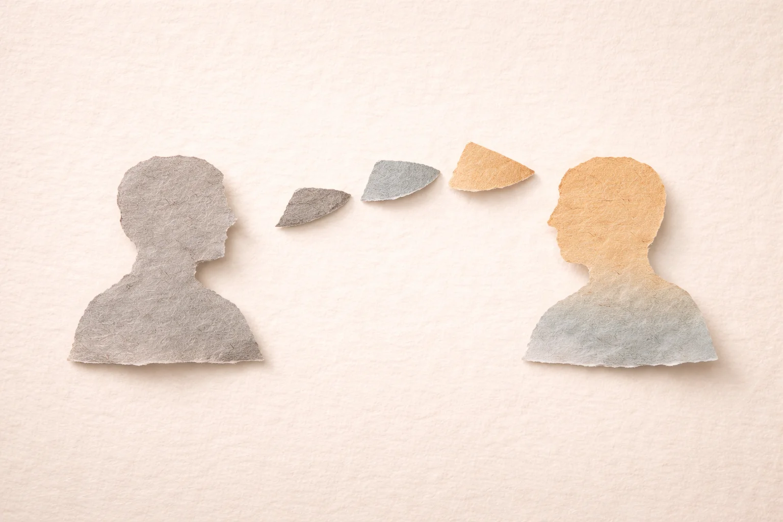 Minimalist torn-paper collage on a warm cream textured background showing two abstract head-and-shoulder silhouettes facing each other, the left in muted grey and the right in a slightly warmer ochre-blue tone, both with rough fibrous edges and soft shadows. Between them, a sequence of small torn-paper shapes flows from left to right in a gentle upward arc, gradually shifting in colour and openness to suggest ideas moving forward through conversation, symbolising a calm, constructive exchange that builds clarity and momentum without relying on words or symbols.