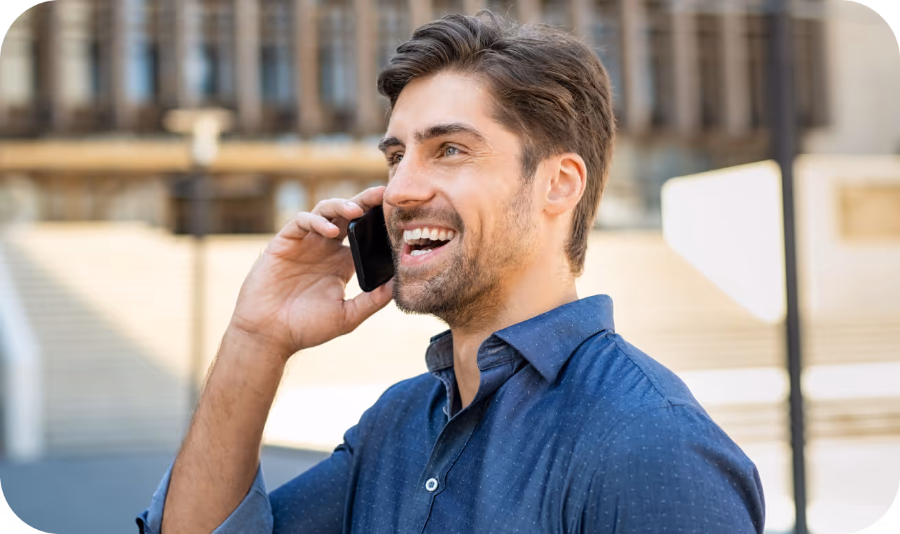 Smiling man in a blue shirt talking on a smartphone outdoors in a city setting.