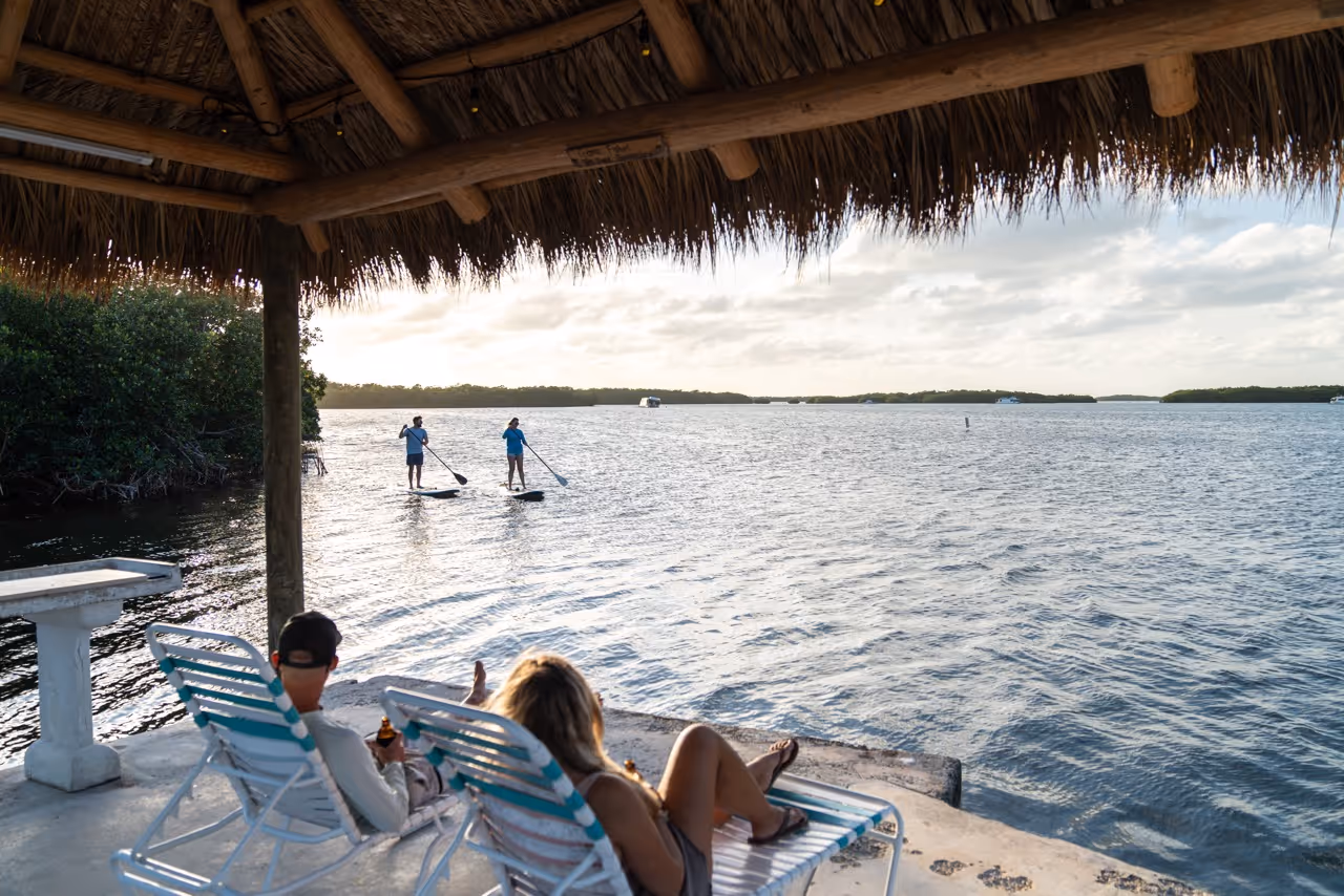 A couple sits under the tiki hut on the water watching two paddleboarders on the Florida Bay.