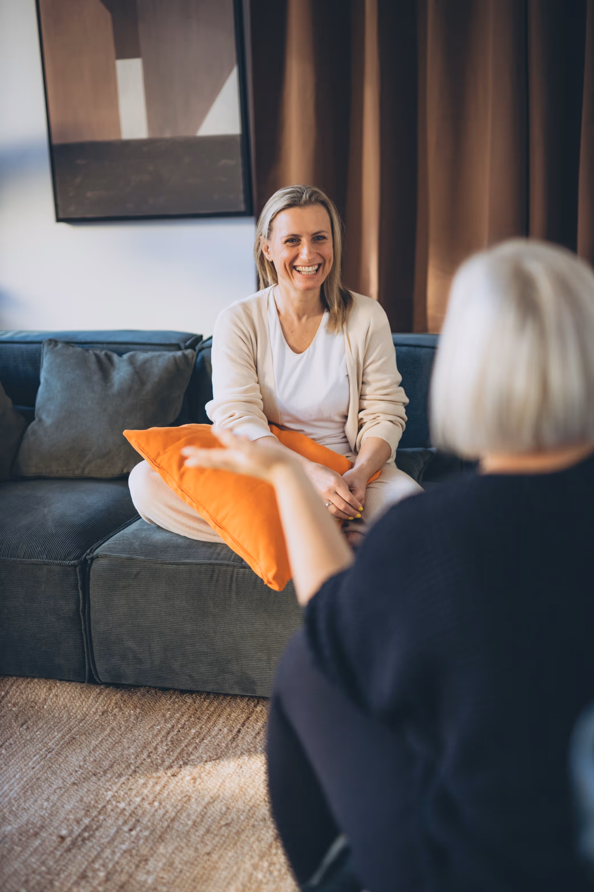 Lächelnde Frau, die ein orangefarbenes Kissen hält und auf einem grauen Sofa sitzt, während sie sich mit Annika Fischer unterhält.