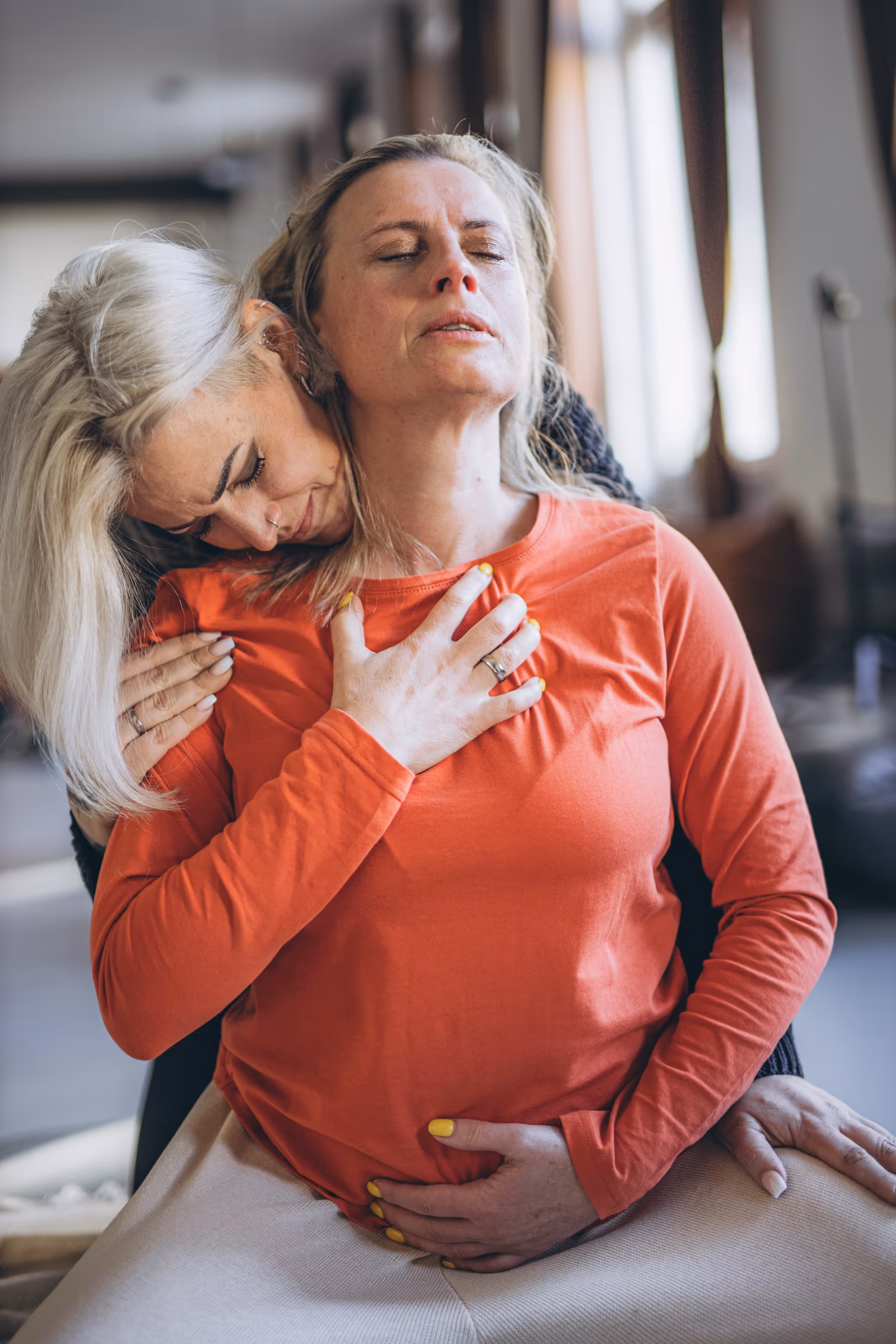Zwei Frauen umarmen sich innig in einem ruhigen, unterstützenden Moment, eine davon trägt ein orangefarbenes Oberteil und hat die Augen geschlossen.