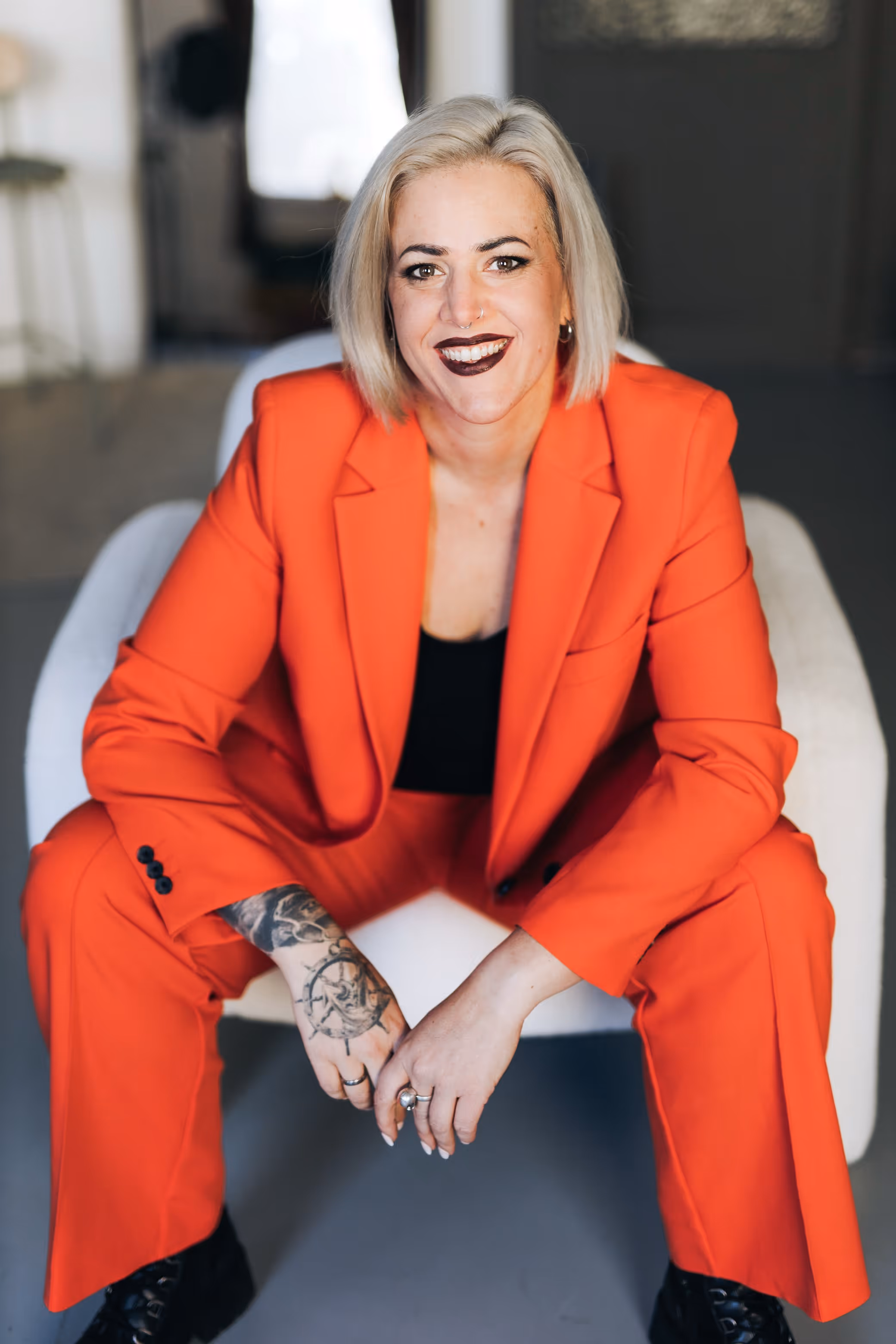 Lächelnde Annika Fischer mit kurzen blonden Haaren, die einen leuchtend orangefarbenen Anzug und ein schwarzes Oberteil trägt, auf einem Stuhl sitzt, die Hände gefaltet hält und ein Tattoo auf ihrem rechten Unterarm zeigt.