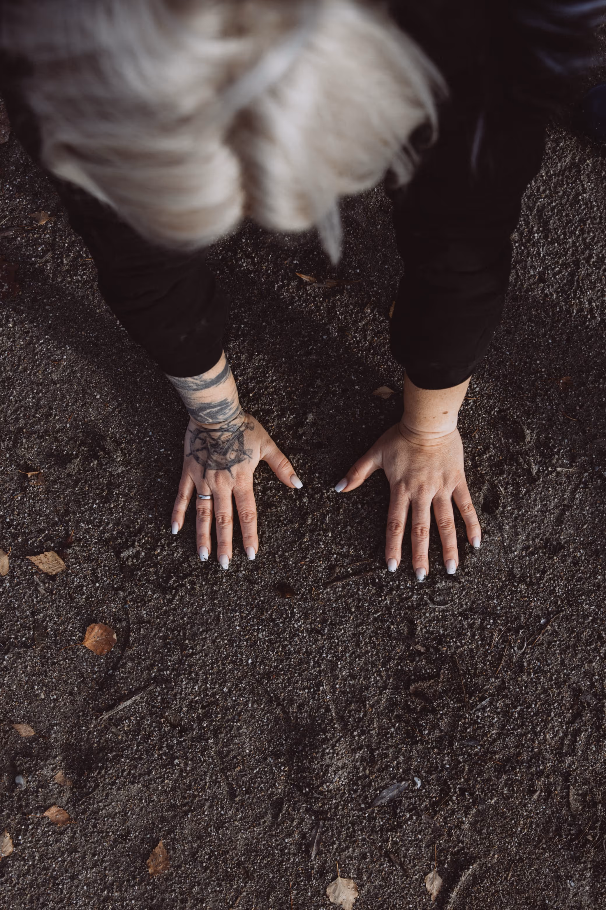 Draufsicht auf eine Person mit blonden Haaren und einer tätowierten linken Hand, die beide Hände auf den mit trockenen Blättern übersäten Boden legt.