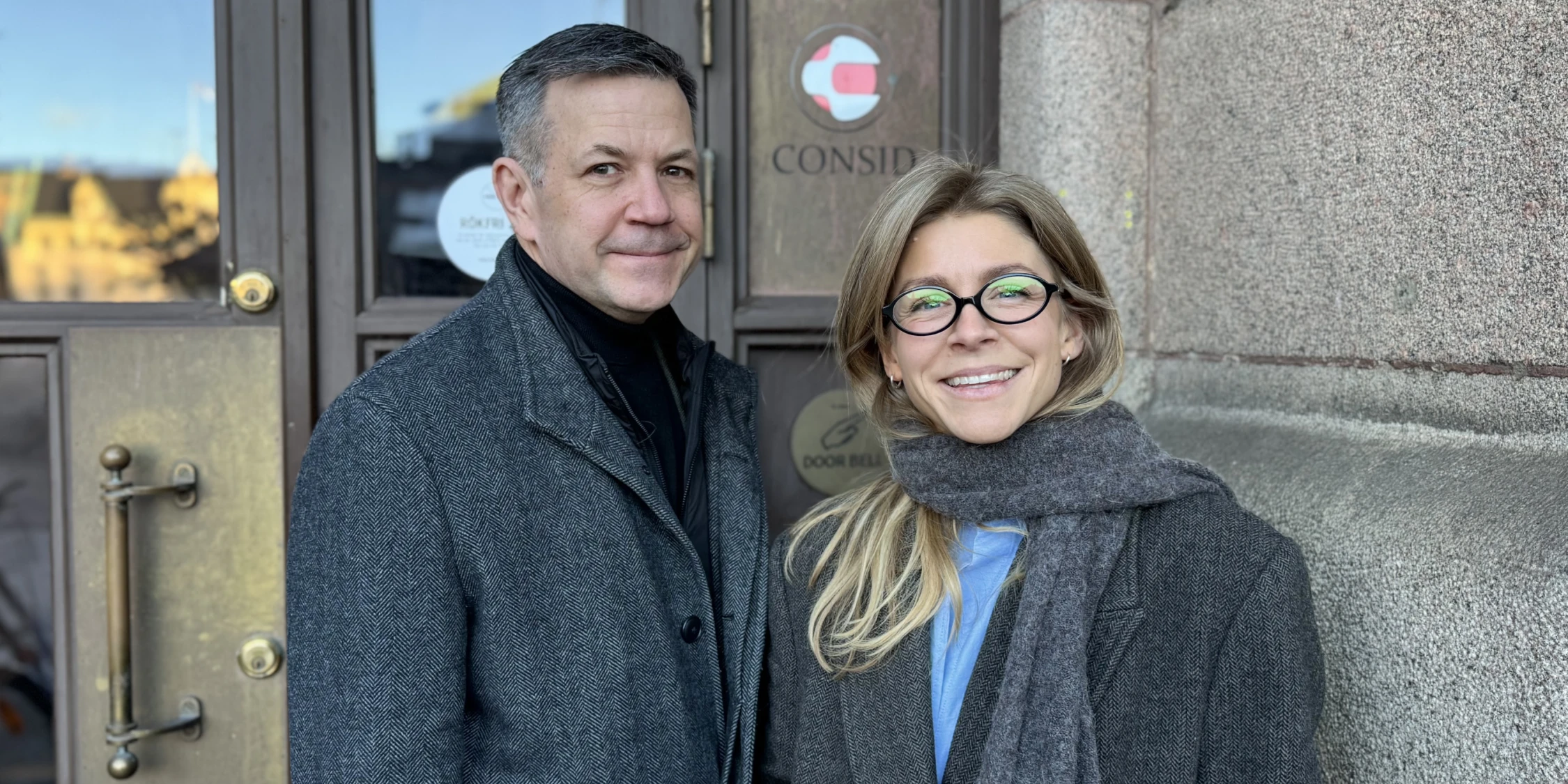 En man och en kvinna står utanför en entré med Consids logotyp på dörren. Båda bär mörka rockar och tittar in mot kameran.