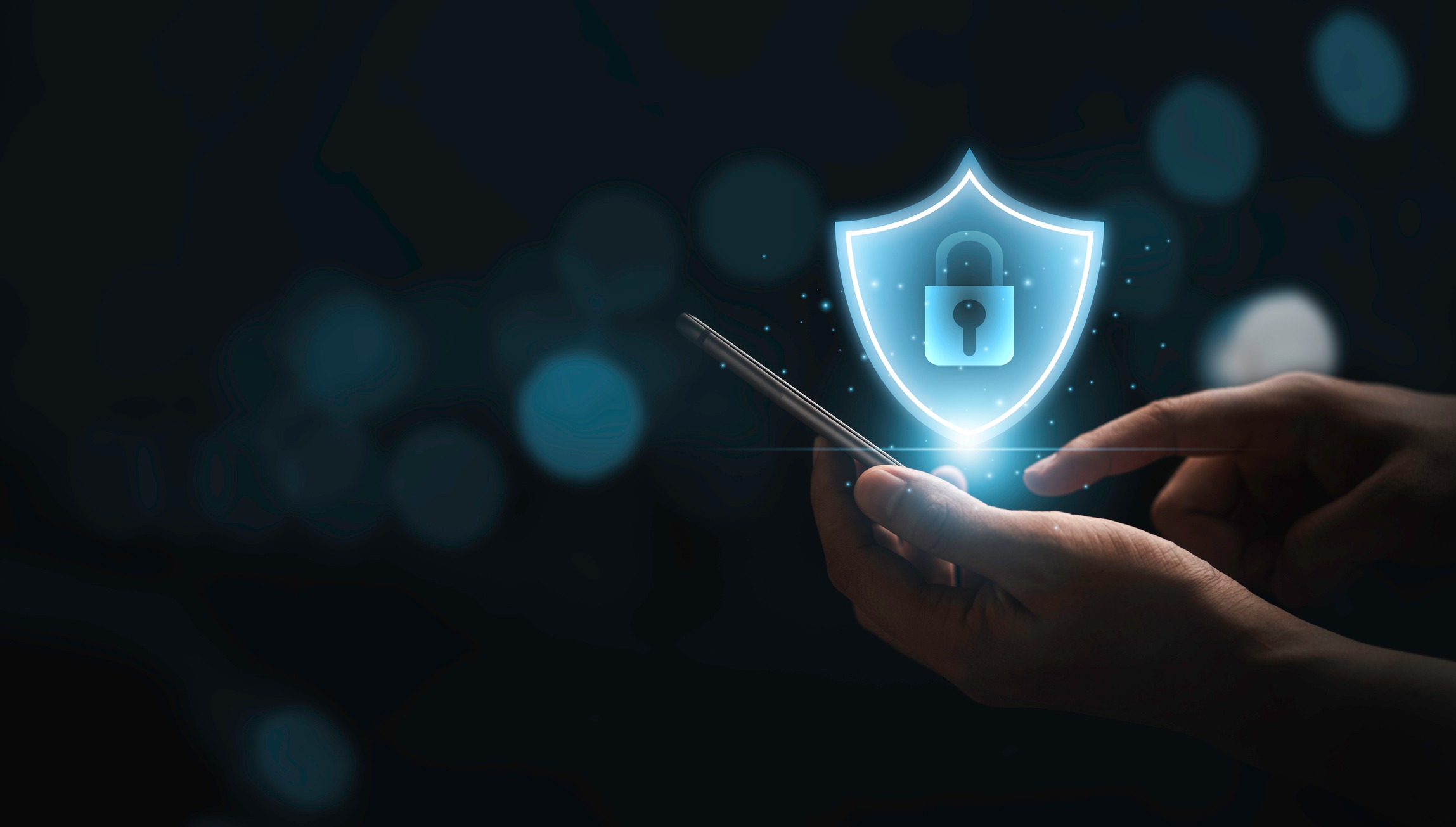A hand holding a smartphone with a glowing blue holographic shield icon displaying a padlock symbol floating above it, set against a dark background with blurred bokeh lights, representing mobile security or cybersecurity protection.