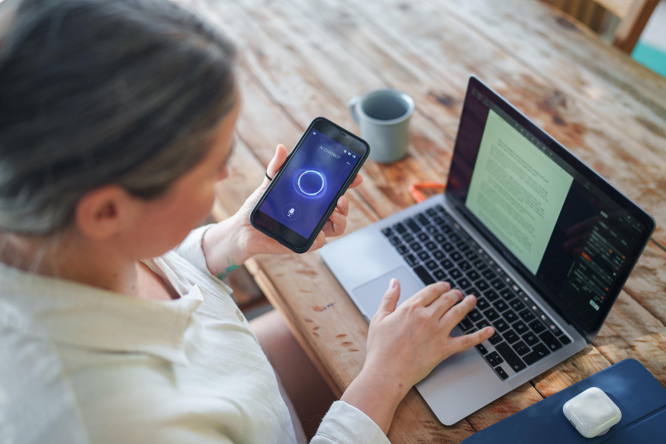 Woman using an AI chatbot application on a smartphone while working on a laptop at a wooden desk