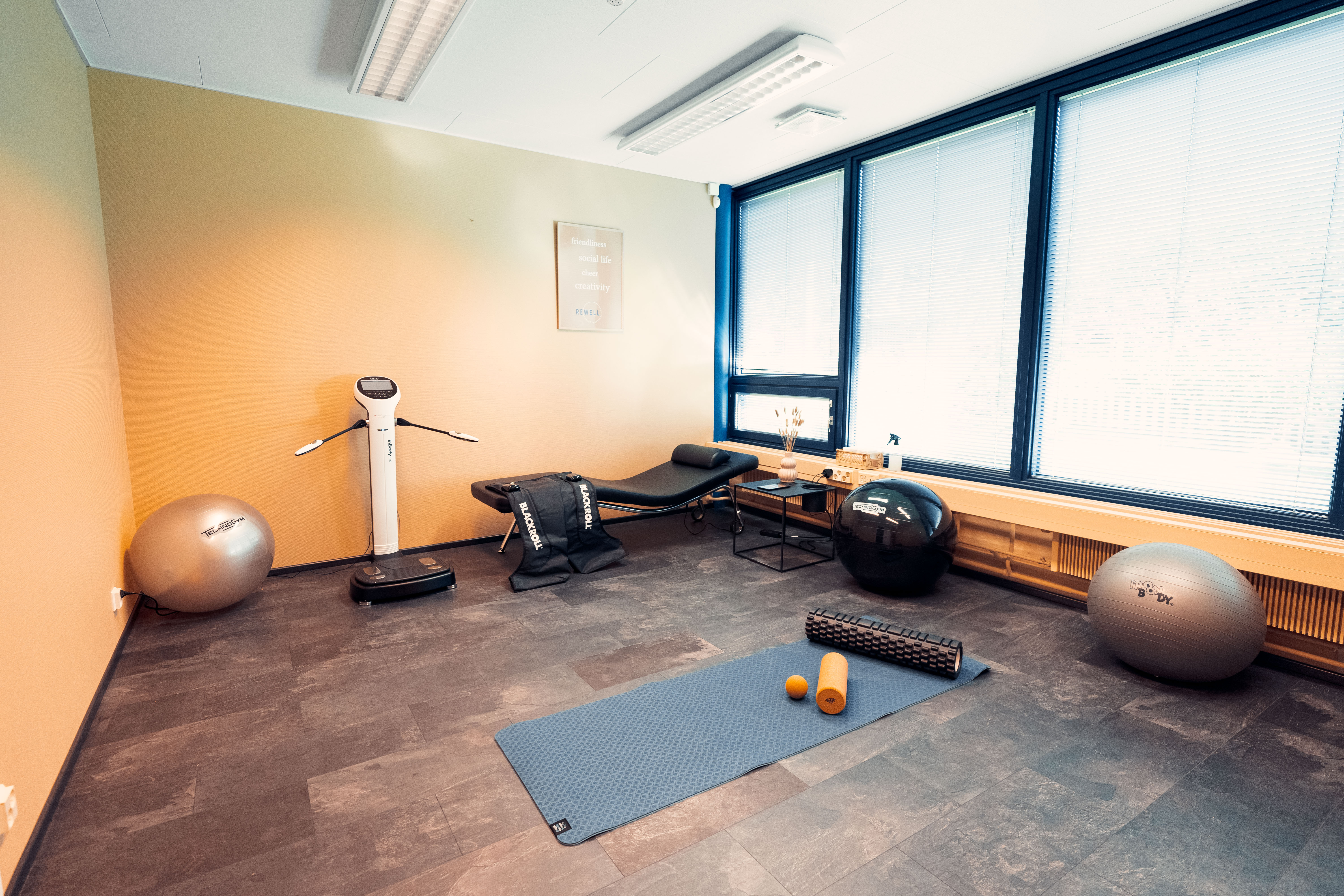 Bright fitness room with exercise balls, a massage table, a vibration plate, a yoga mat with foam rollers and massage balls, and large windows with blinds.