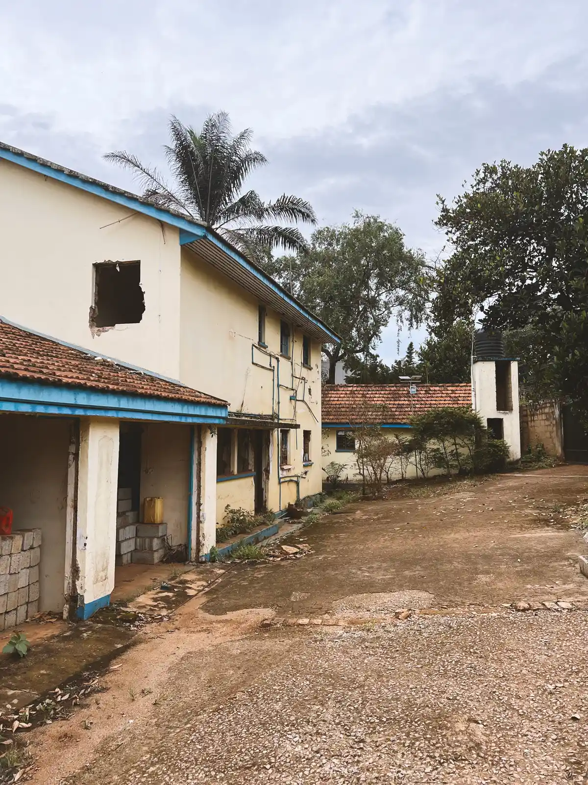 View of a large beige building with blue trim, tiled roofs, an unfinished or damaged window, and a concrete courtyard surrounded by trees. The Ark Preschool Kampala Uganda.