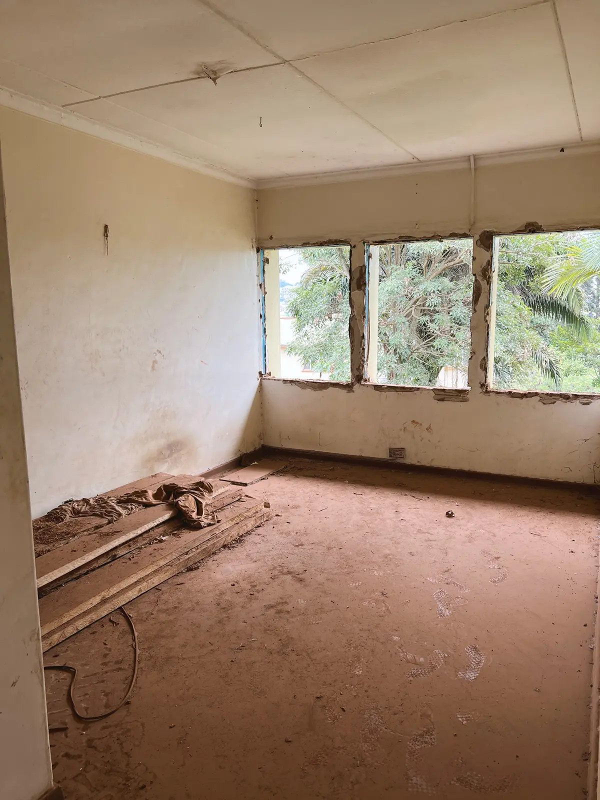 Empty room under construction with bare walls, dirt floor, wooden planks stacked in a corner, and three open windows showing green trees outside. The Ark Preschool Kampala Uganda.