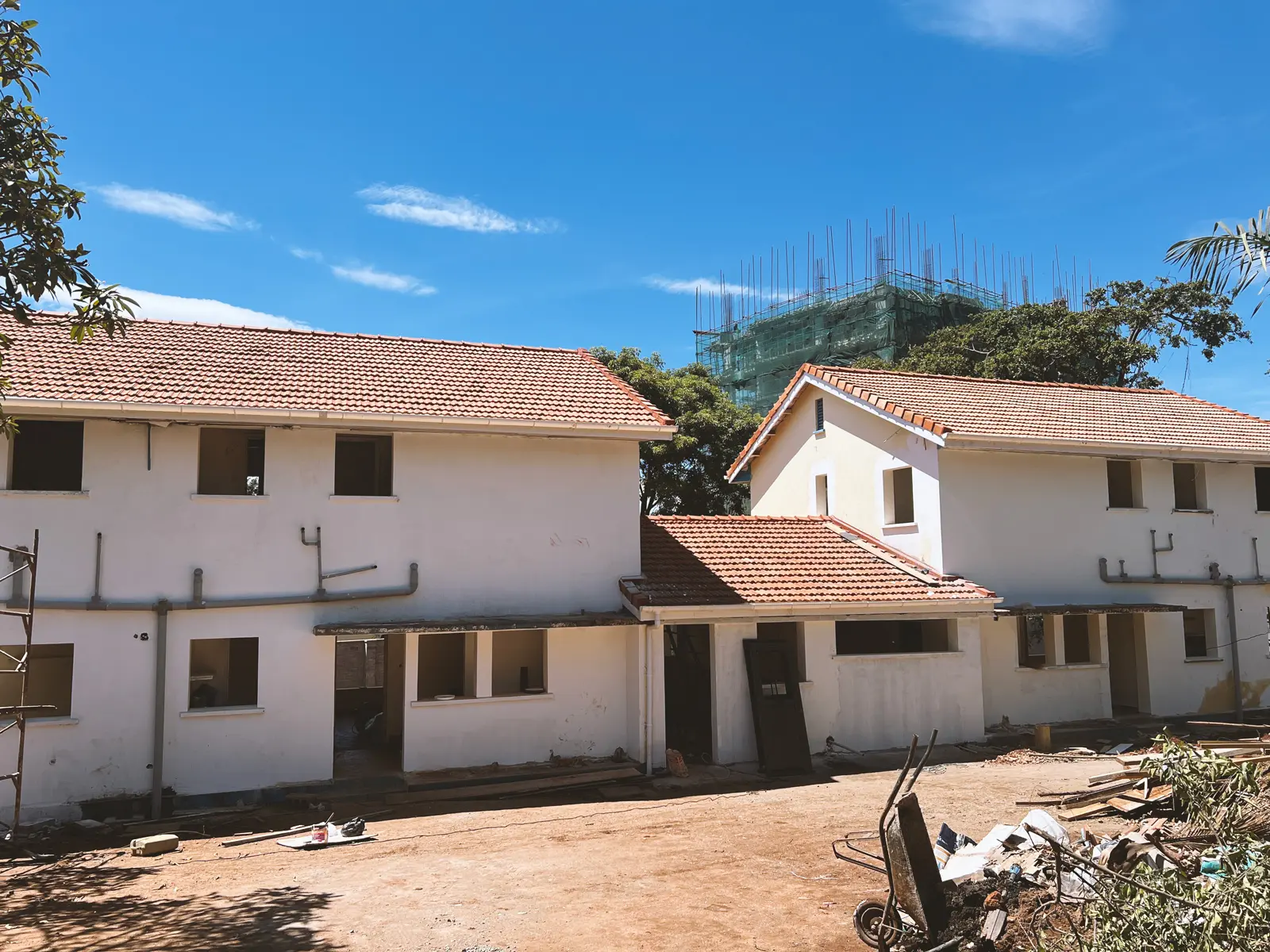 Two white partially constructed buildings with red-tiled roofs under a clear blue sky, surrounded by construction materials and trees. The Ark Preschool Kampala Uganda.