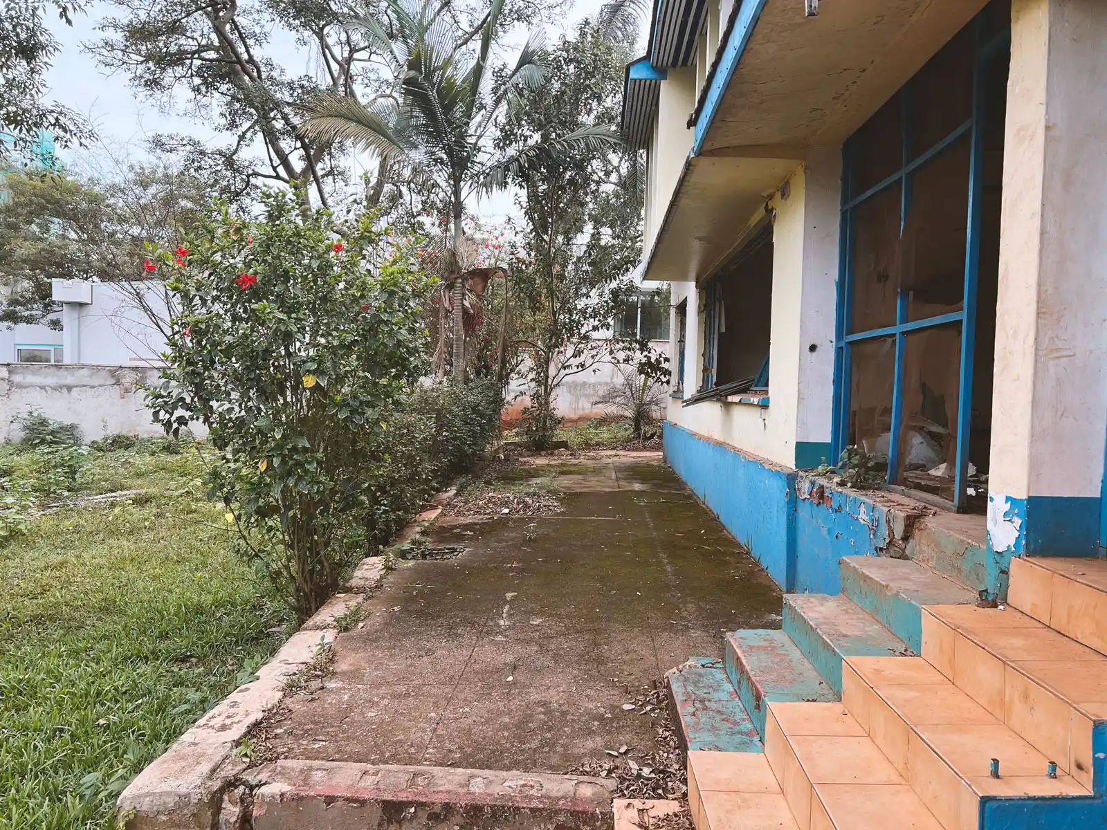Exterior walkway beside an old, weathered building with chipping blue paint and a bush with red flowers on the left. The Ark Preschool Kampala Uganda.