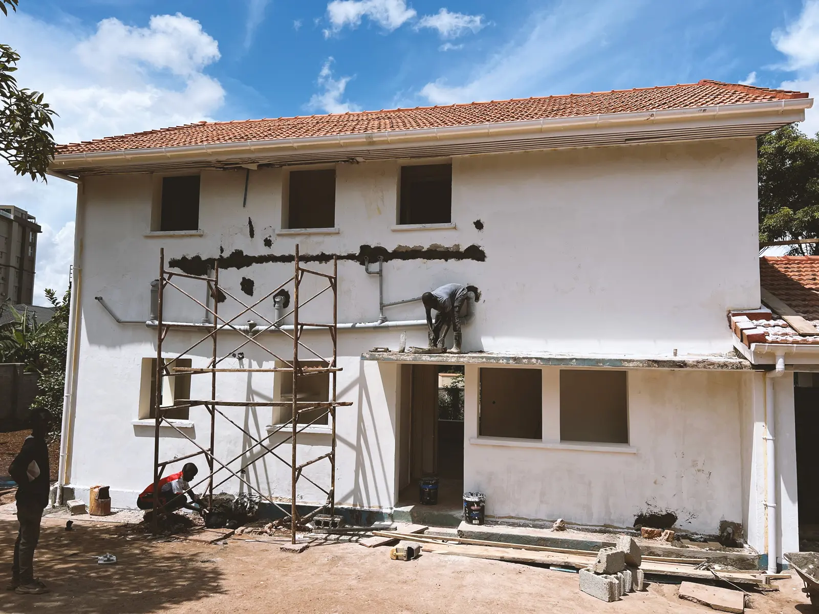 Two workers repairing and plastering the white exterior wall of a two-story house with a red tiled roof under a blue sky. The Ark Preschool Kampala Uganda.