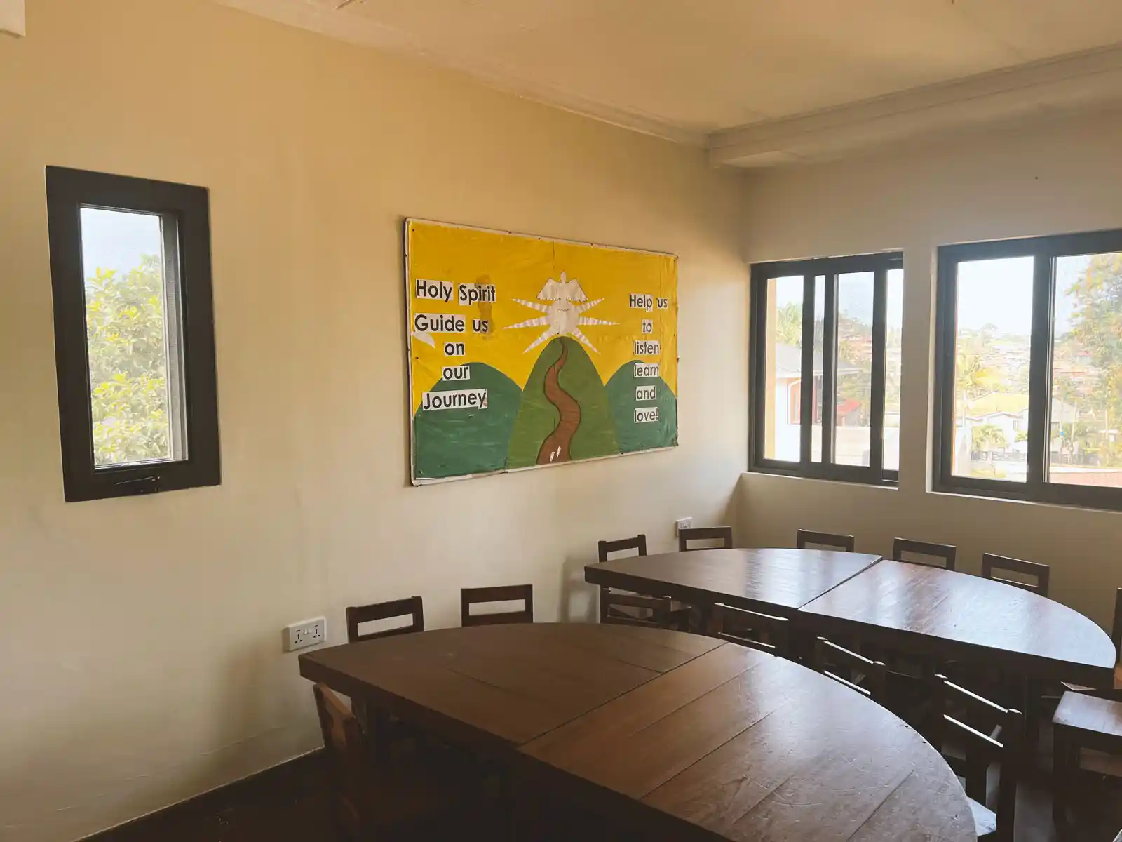 Classroom with wooden tables and chairs arranged by windows, featuring a yellow bulletin board with a religious message and artwork on the wall. The Ark Preschool Kampala Uganda.