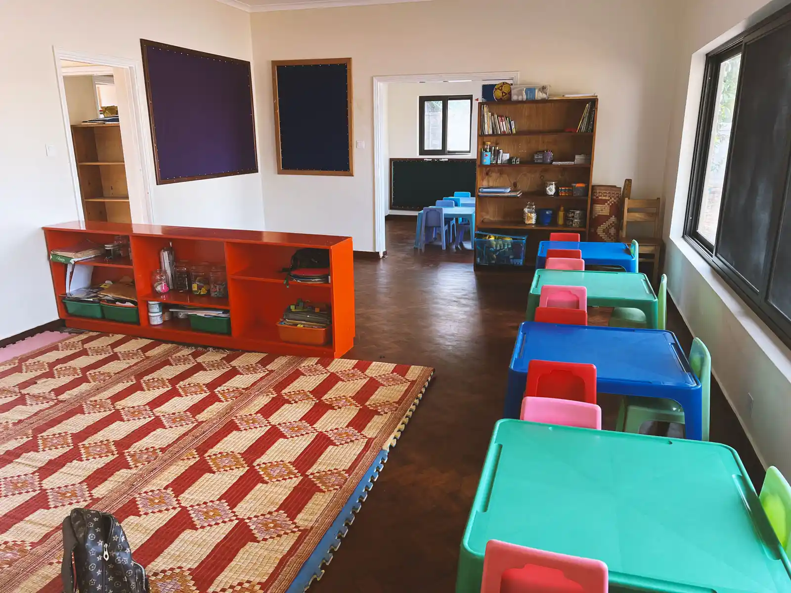 Empty preschool classroom with colorful small tables and chairs, shelves with supplies, and a patterned floor mat. The Ark Preschool Kampala Uganda.