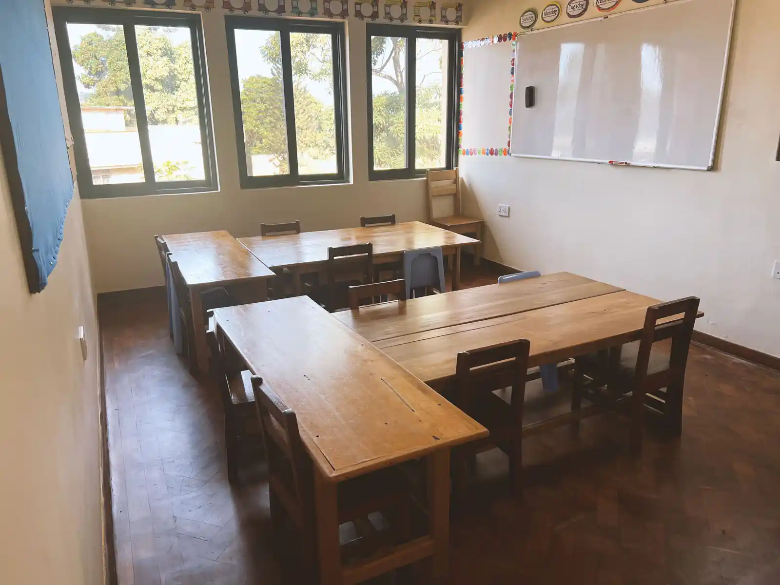 Empty classroom with wooden tables and chairs arranged under large windows and a whiteboard on the wall. The Ark Preschool Kampala Uganda.