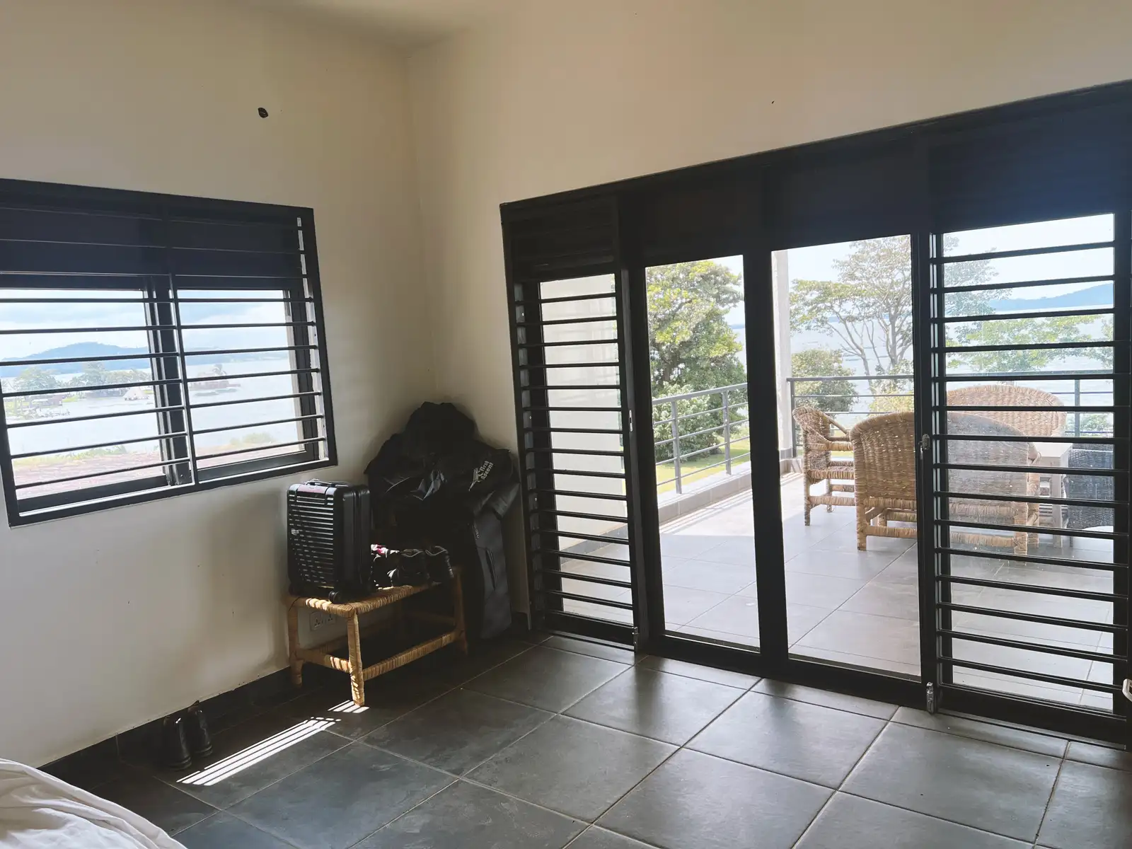 Minimalist room with tiled floor, black window shutters, small rattan stool with luggage, and sliding doors opening to a balcony with wicker chairs and a scenic view. Busalaba Residential Remodel in Kampala by Elevate Holdings Limited.