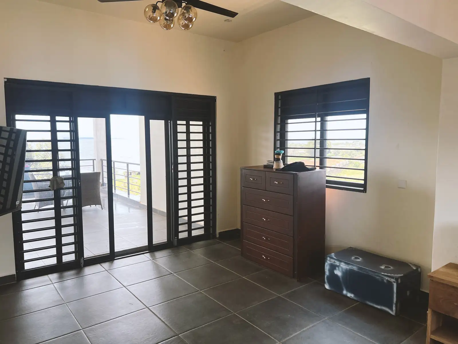 Room with large tiled floor, dark wooden dresser, window with black bars, and sliding glass door opening to a balcony overlooking water. Busalaba Residential Remodel in Kampala by Elevate Holdings Limited.