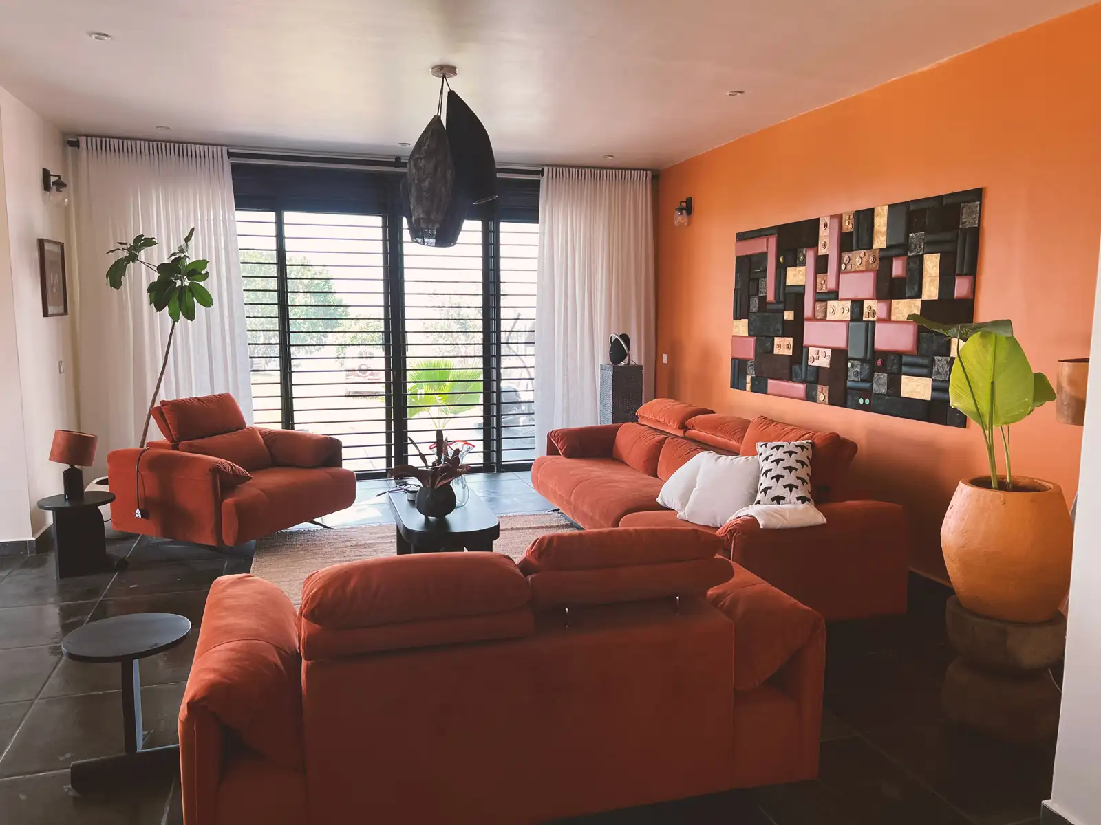 Modern living room with orange sofas, armchair, black coffee table, large window with white curtains, orange accent wall, and decorative wall art. Busalaba Residential Remodel in Kampala by Elevate Holdings Limited.