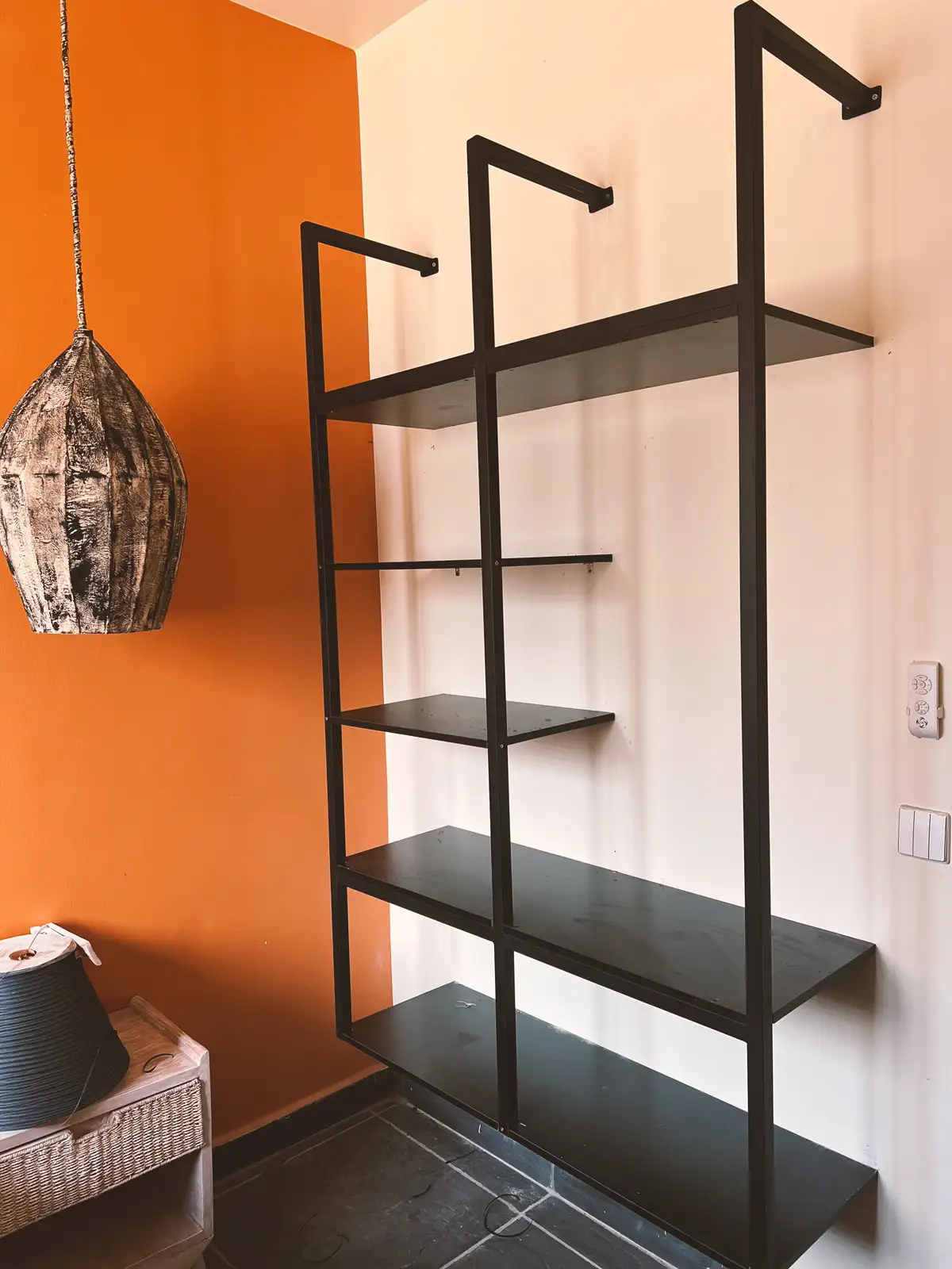Black metal shelving unit mounted on a wall with orange and white paint, next to a woven basket on a wooden table and a hanging rustic pendant light. Busalaba Residential Remodel in Kampala by Elevate Holdings Limited.