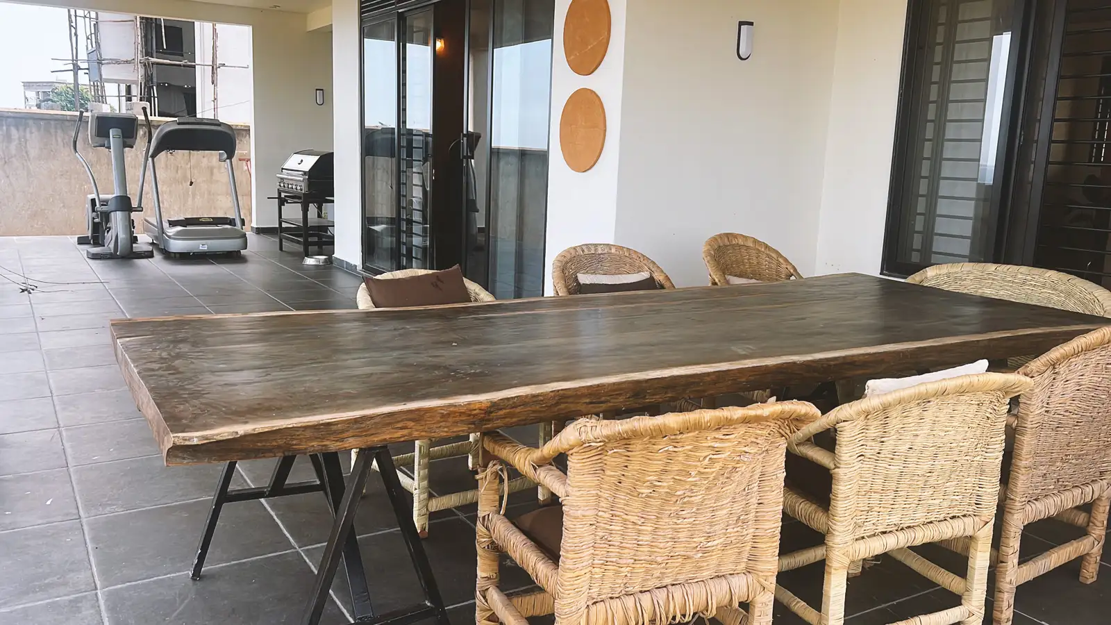 Outdoor patio with a large wooden dining table surrounded by wicker chairs, and exercise equipment in the background. Busalaba Residential Remodel in Kampala by Elevate Holdings Limited.