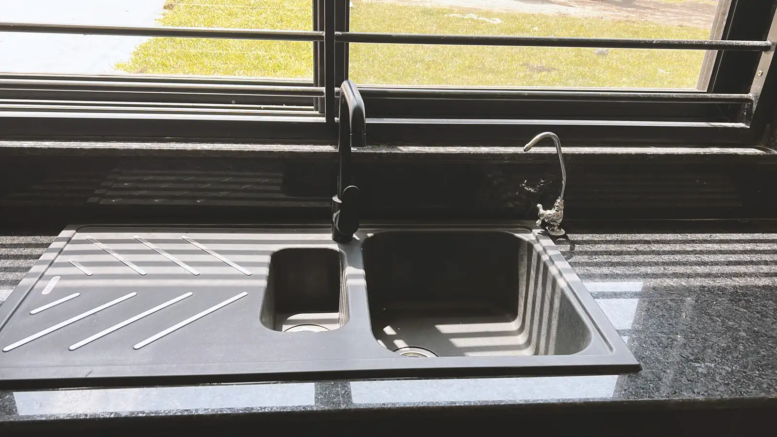 Modern double kitchen sink with black and silver faucets on black granite countertop, sunlight casting shadows through window blinds. Busalaba Residential Remodel in Kampala by Elevate Holdings Limited.