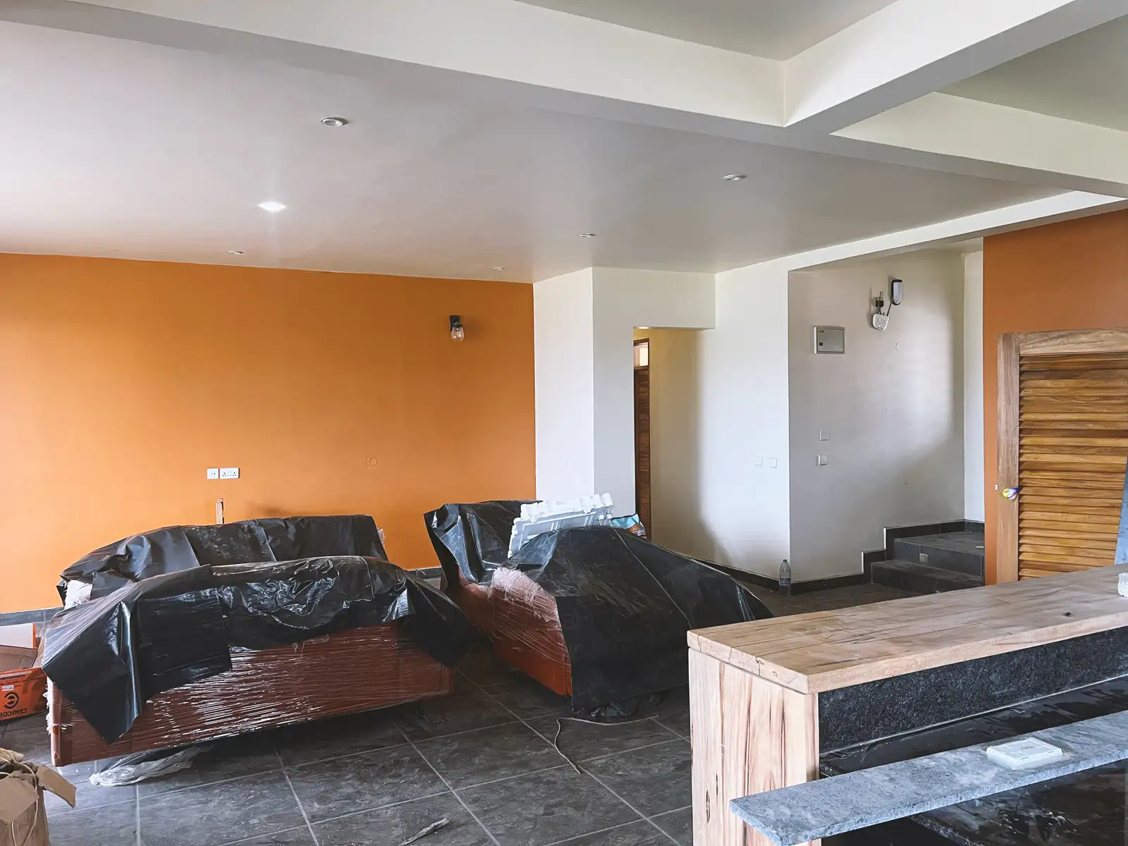 Living room under renovation with orange and white walls, covered furniture, tiled floor, and unfinished countertop.