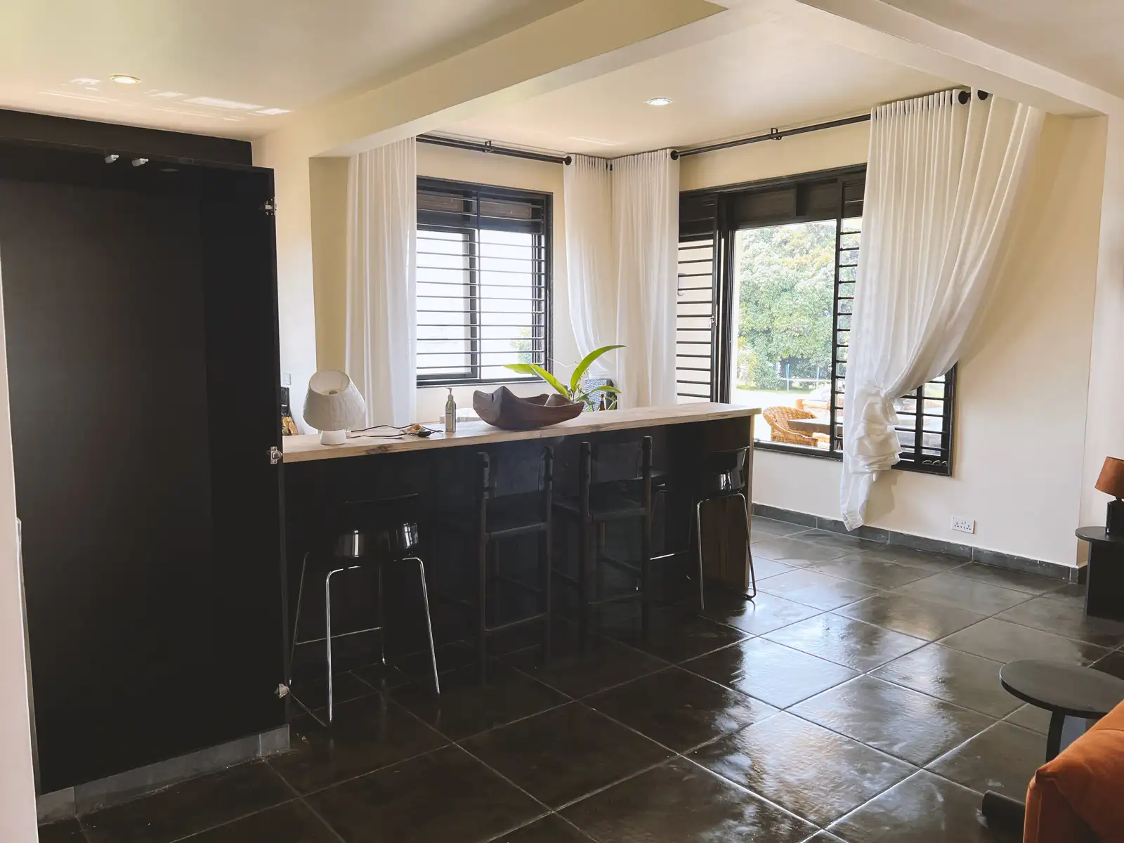 Modern room with black tile floor, long counter with three black chairs, large windows with white curtains, and a view of outdoor greenery.