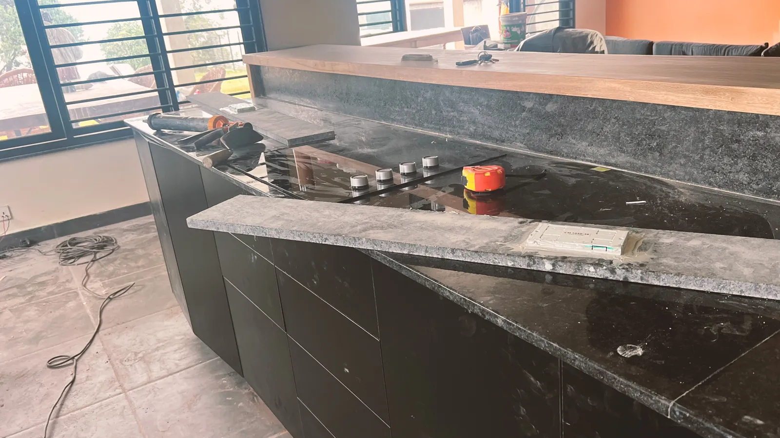 Kitchen countertop under construction with black granite surfaces, silver knobs, measuring tape, tools, and loose wiring on the floor.