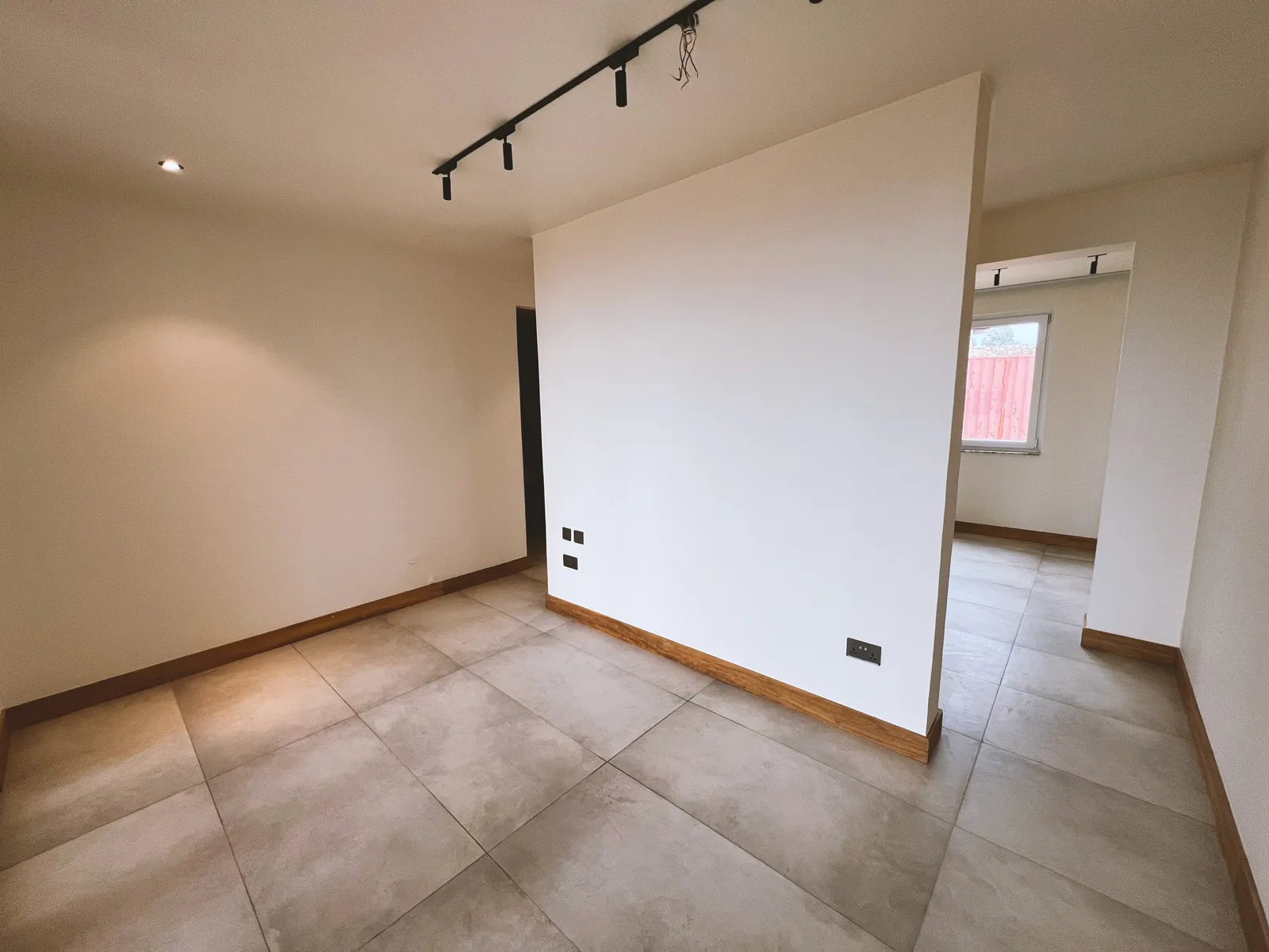 Empty room with beige tiled floor, white walls with wooden baseboards, a window looking outside, and ceiling track lighting. Sonde Residential Remodel by Elevate Holdings Limited.