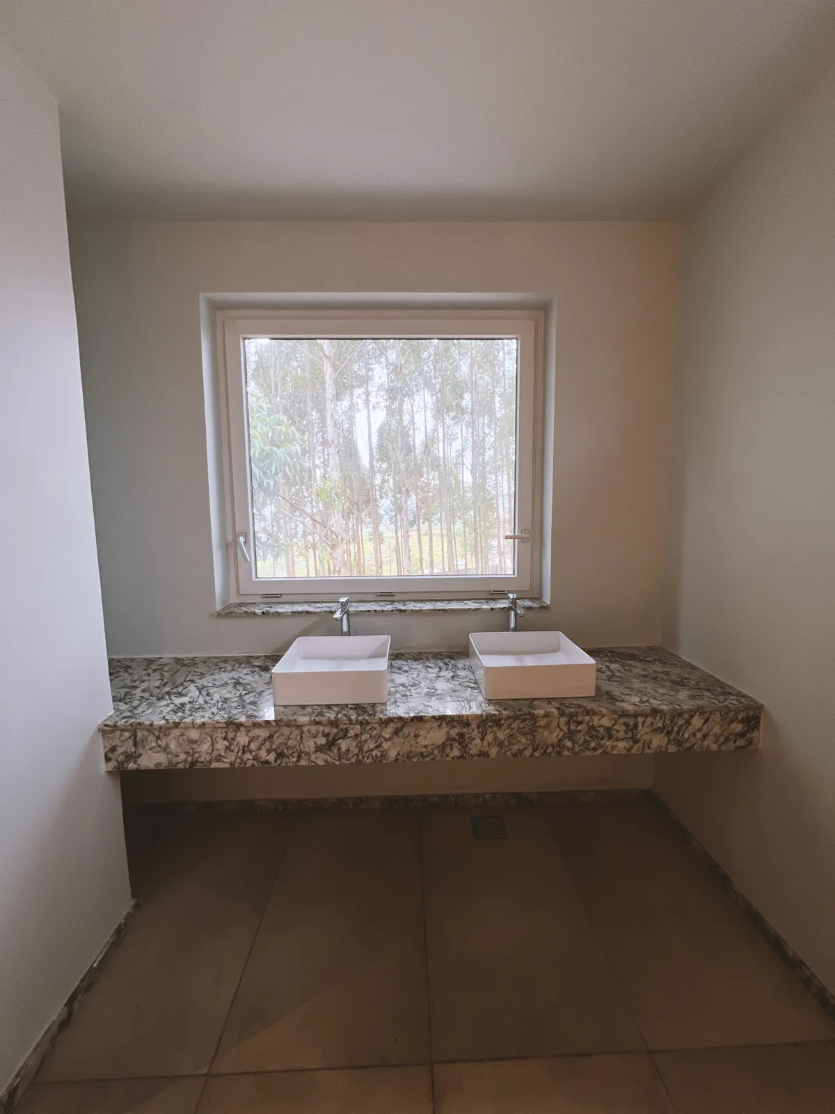 Minimal bathroom with two square white sinks on a marble countertop under a window showing outdoor trees. Sonde Residential Remodel by Elevate Holdings Limited.