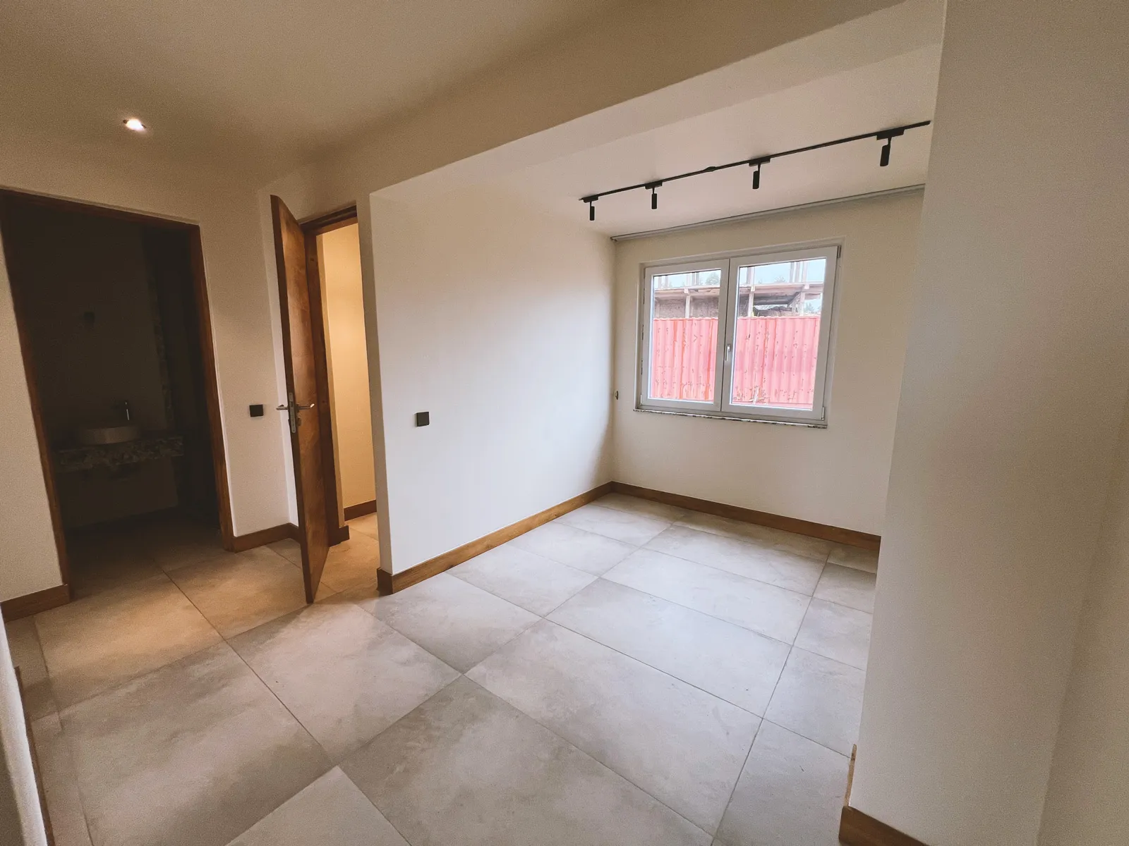 Empty room with beige tiled floor, white walls, a window with a view of a red fence, and two wooden doors, one partially open. Sonde Residential Remodel by Elevate Holdings Limited.