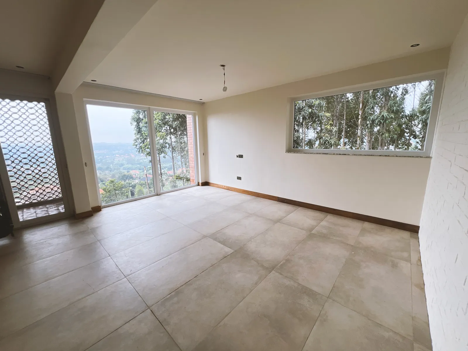 Empty room with large beige floor tiles, sliding glass doors with a view of trees and distant landscape, and a rectangular window showing tree foliage. Sonde Residential Remodel by Elevate Holdings Limited.