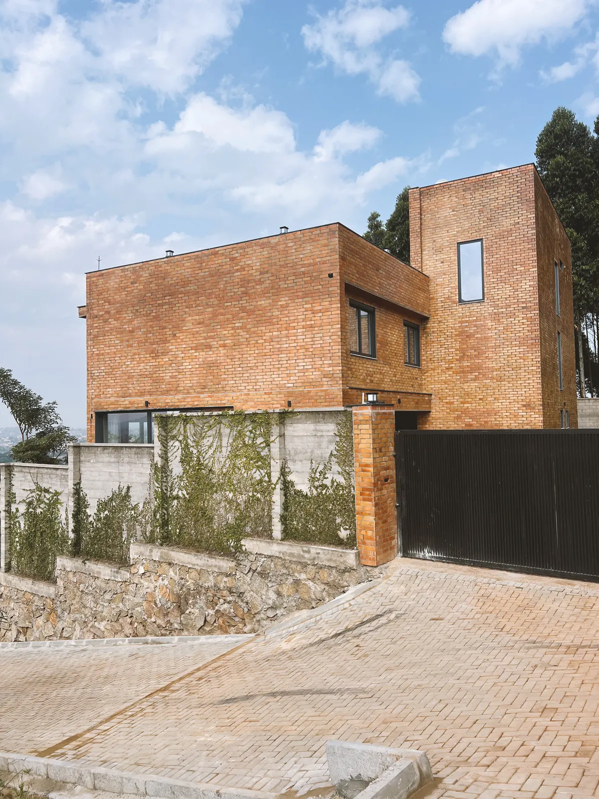 Modern brick house with large windows, concrete and stone walls with climbing plants, and a black gate under a partly cloudy sky. Sonde Residential Remodel by Elevate Holdings Limited.
