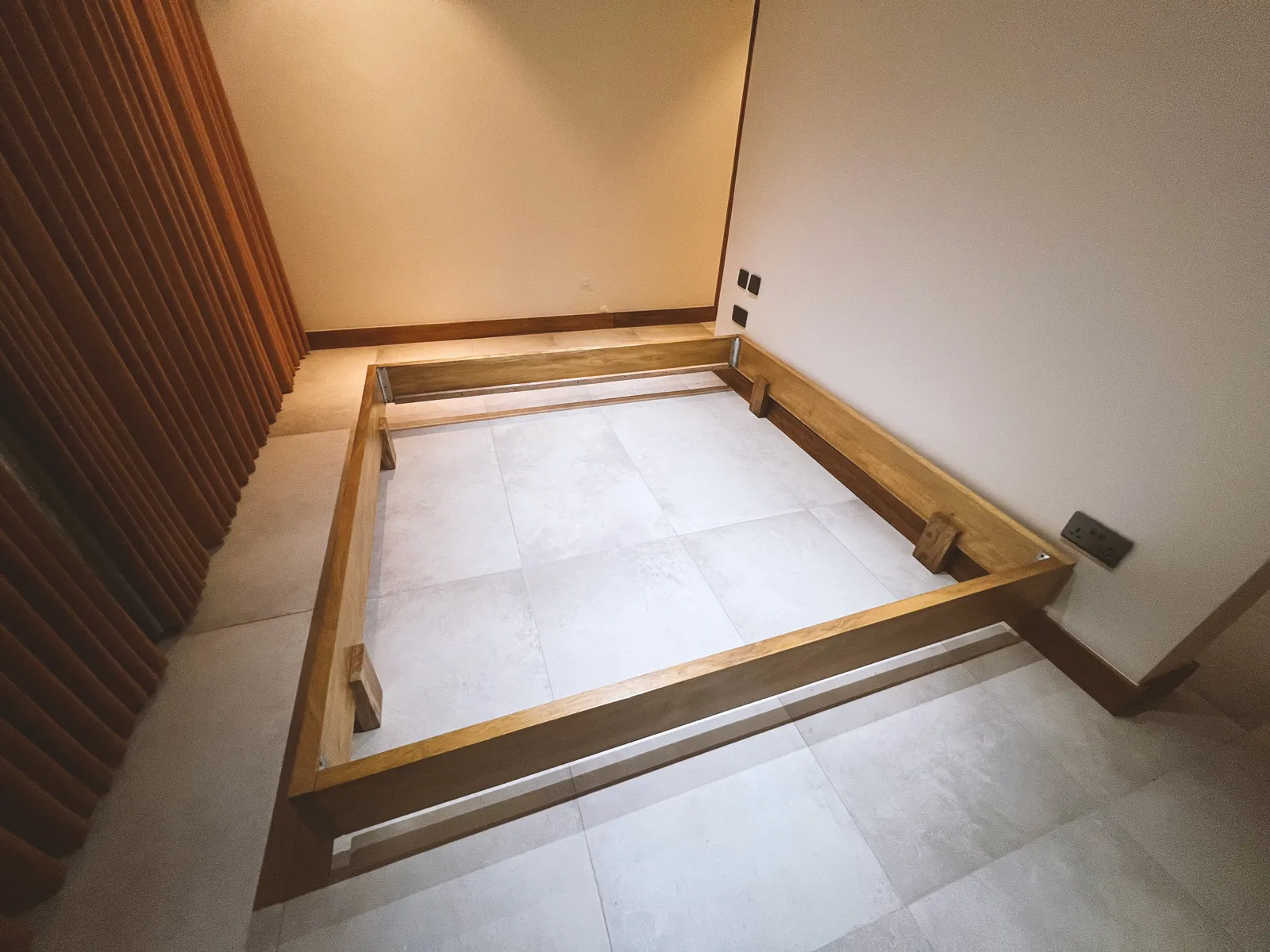 Empty wooden bed frame placed on tiled floor in a corner of a room with beige walls and brown curtains. Sonde Residential Remodel by Elevate Holdings Limited.