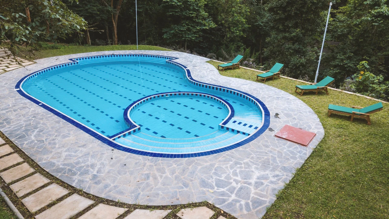 Outdoor swimming pool with an attached circular shallow pool surrounded by stone tiles and green lounge chairs on grassy area. Kalinzu Eco Lodge Construction Project by Elevate Holdings Limited.