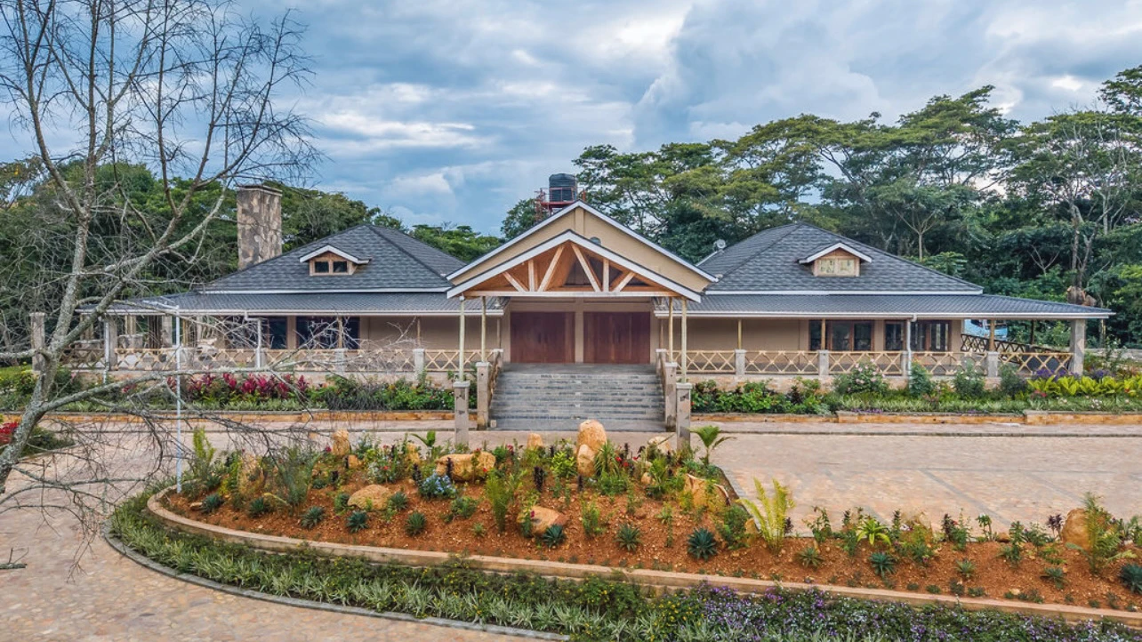 Front view of a large eco-lodge with a gray roof, wooden double doors, surrounded by landscaped gardens and trees under a cloudy sky. Kalinzu Eco Lodge Construction Project by Elevate Holdings Limited.