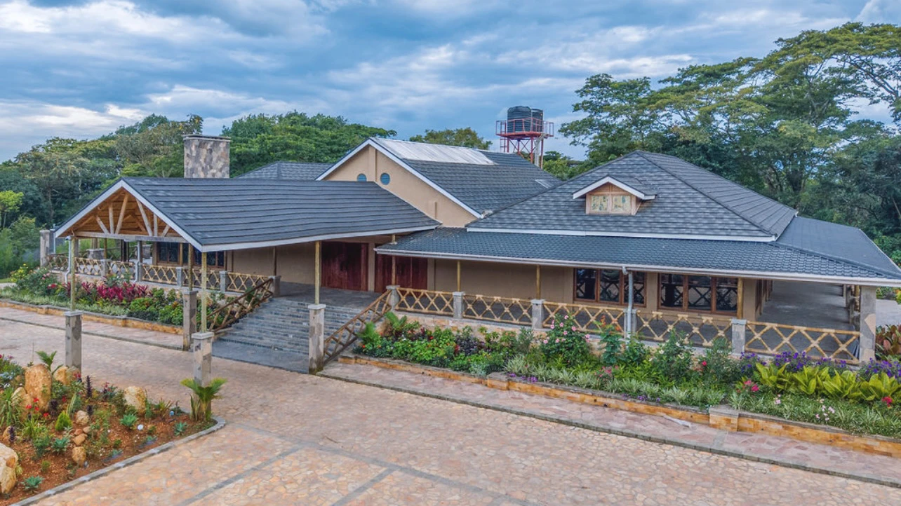 Single-story building with dark gray sloped roofs, steps leading to the entrance, surrounded by flower beds and trees under a cloudy sky. Kalinzu Eco Lodge Construction Project by Elevate Holdings Limited.