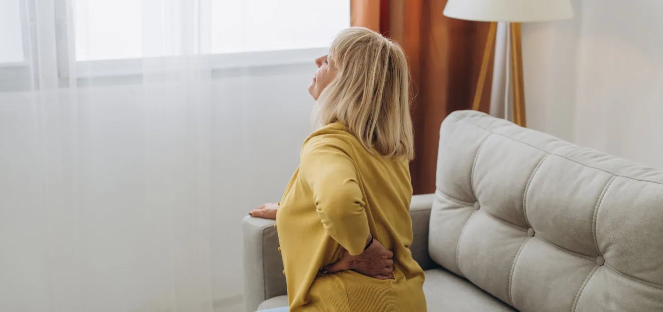 Symptom bei Bandscheibenvorfällen - Frau sitzt auf der Couch und hat akute Schmerzen und Lähmungserscheinungen durch einen Bandscheibenvorfall