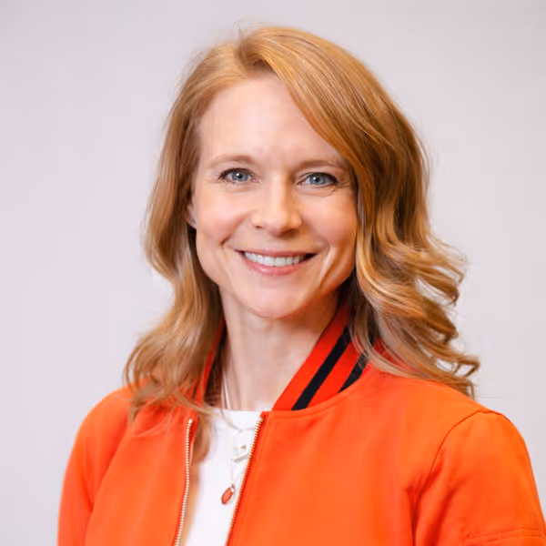Smiling woman with shoulder-length wavy blonde hair wearing an orange jacket with black stripes on the collar.