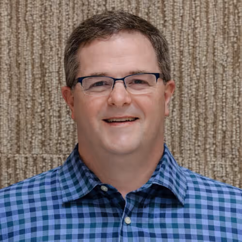 Tom Shanahan. Smiling man wearing glasses and a blue checkered shirt against a textured beige background.